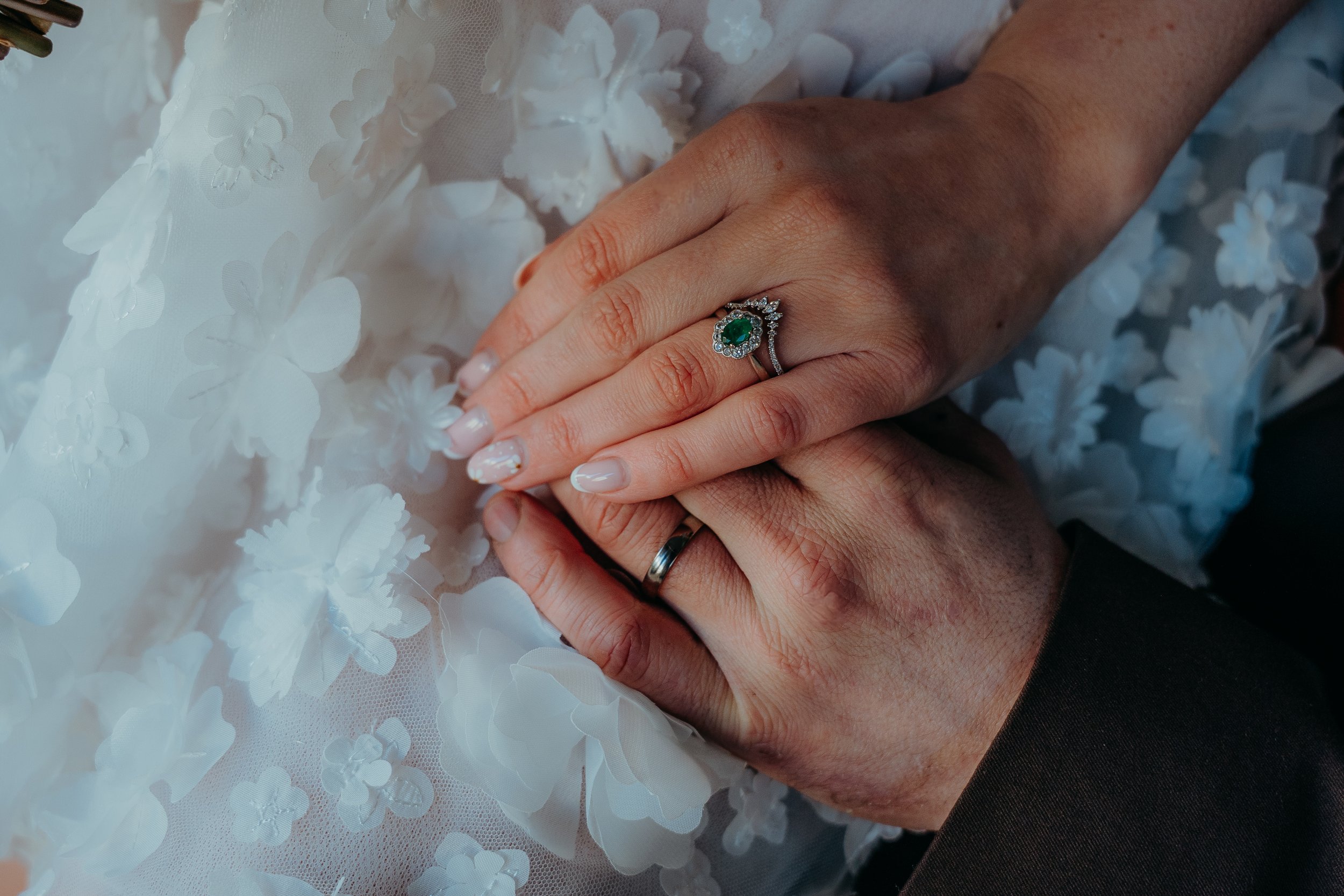 Close-up of a person's hand with a ring featuring a green gemstone surrounded by small diamonds, resting on a white floral lace fabric. Iris willow photography bar 131 Cheltenham