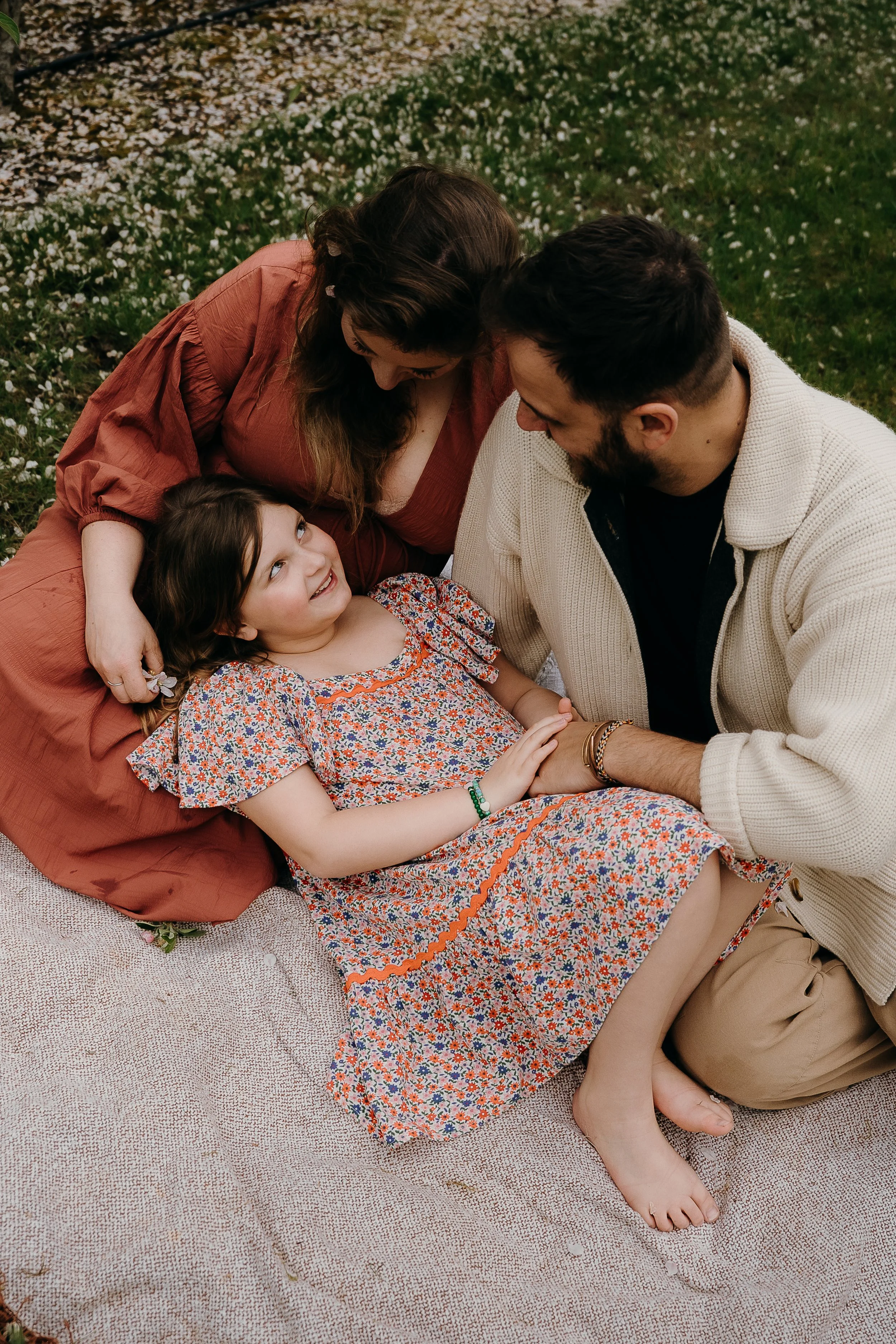Family lying on a blanket outdoors, with a woman and a man gazing at a young girl with long curly brown hair, wearing a floral dress, who is smiling and looking up at them.