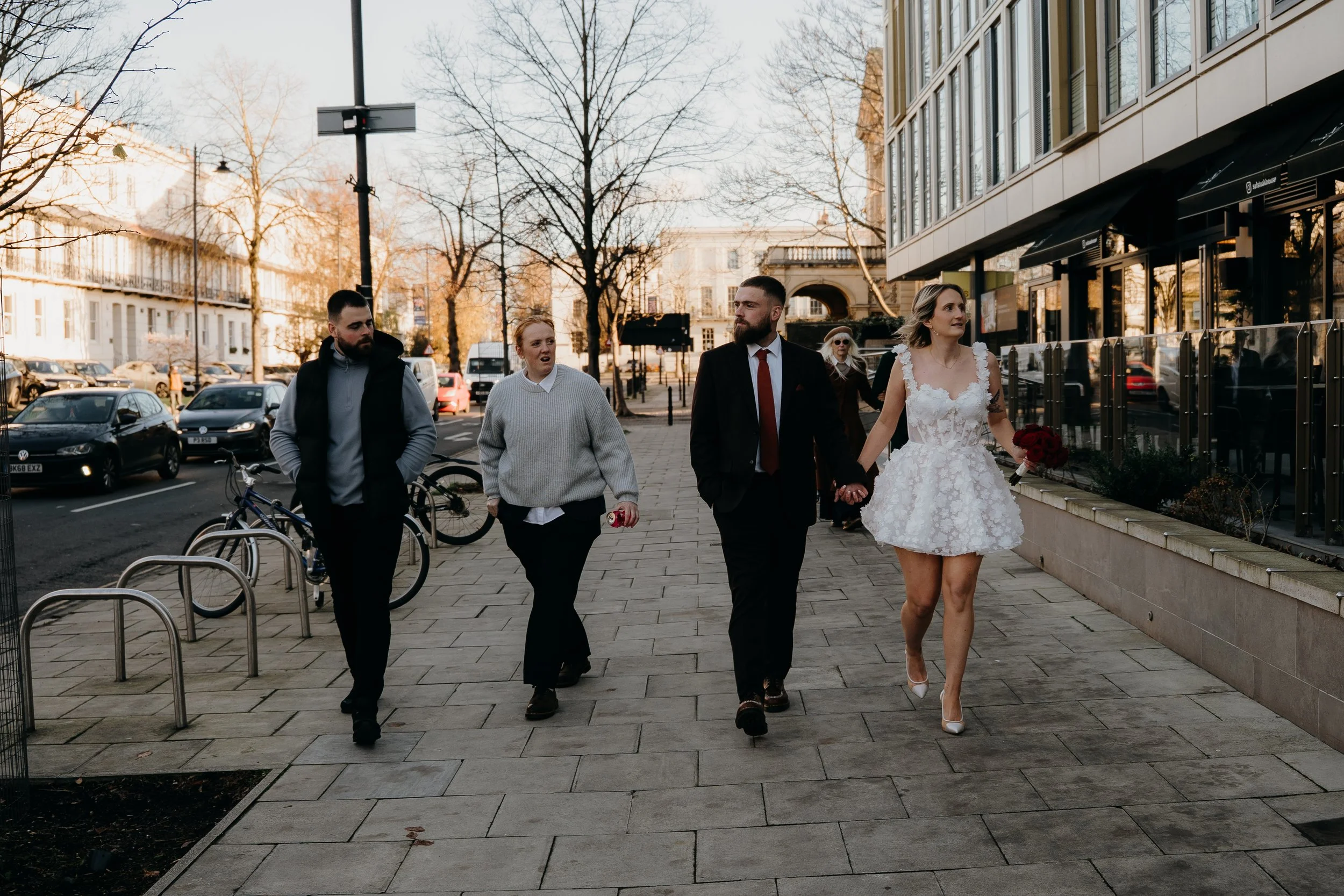 A bride in a white lace wedding dress holding a bouquet of red roses walking with three guests on a city sidewalk in the late afternoon, with parked cars, trees, and buildings in the background.