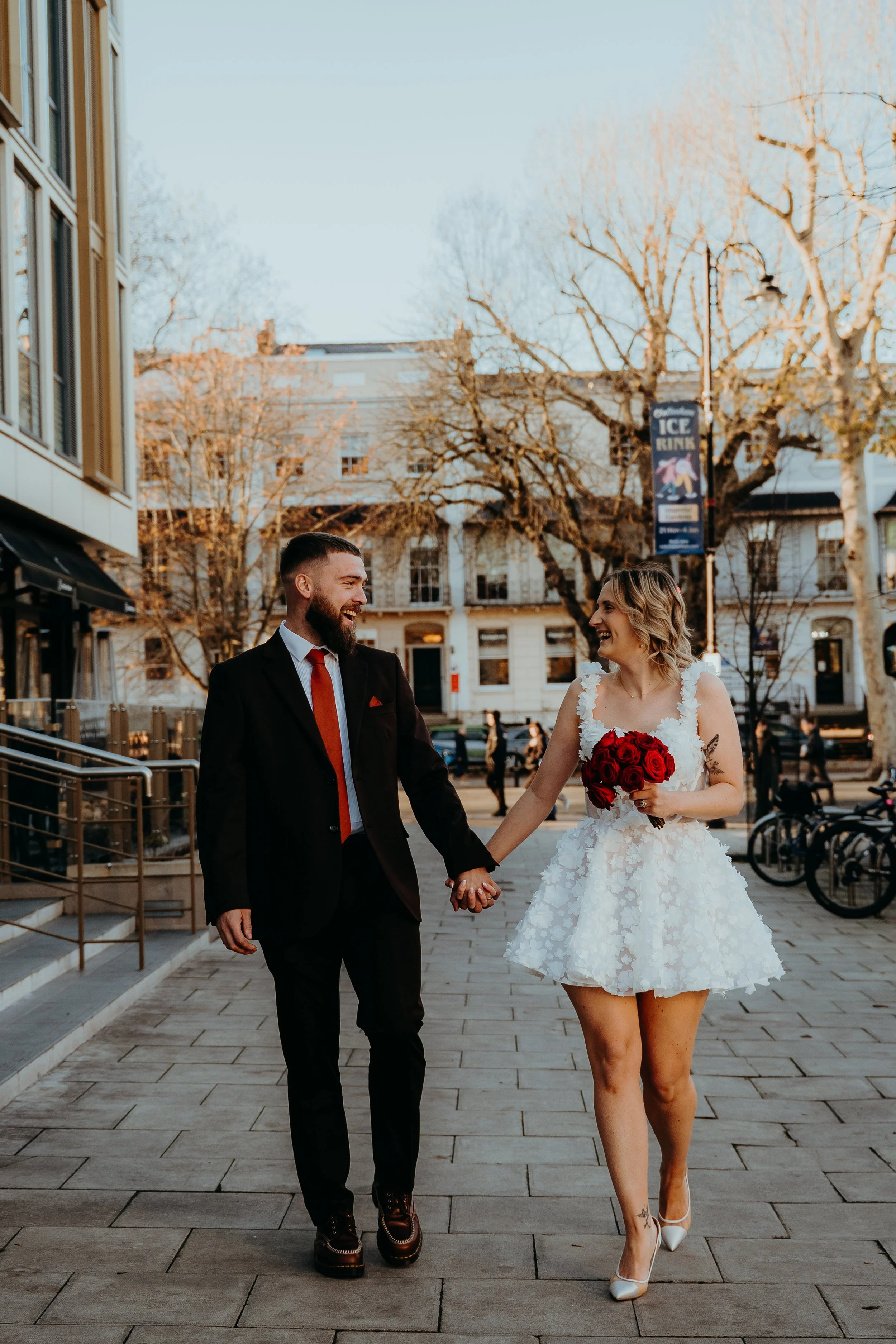 A couple in wedding attire walking hand in hand outdoors, with the woman holding a bouquet of red roses, smiling and looking at each other, in an urban setting with buildings, trees, and bicycles in the background. Iris willow photography bar 131 Che