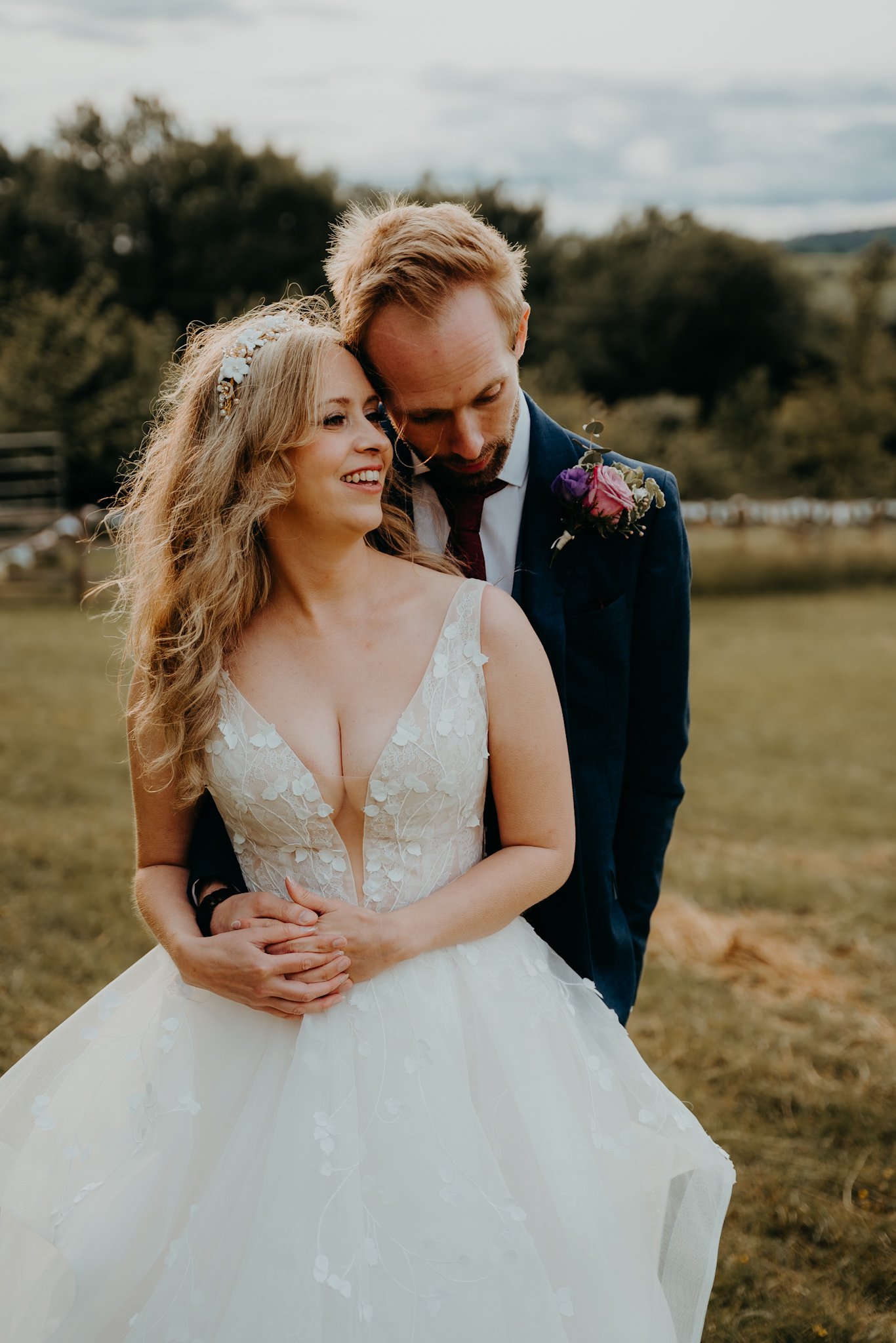 A bride and groom standing outdoors on a cloudy day, embracing each other happily, with green trees and a fence in the background.