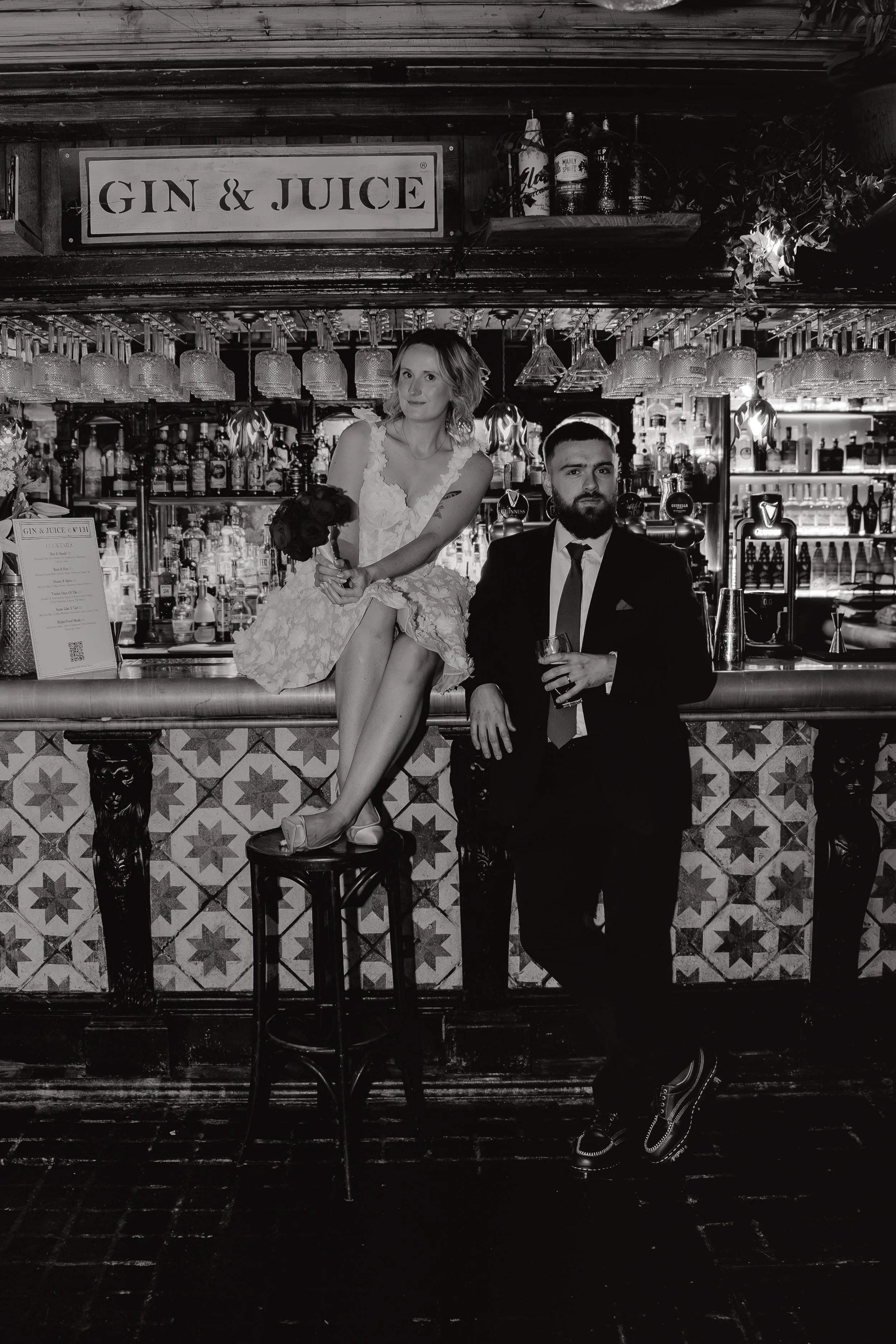 A black and white photo of a bar scene with a Bride and groom holding cocktails at bar 131 Cheltenham. The background has shelves with bottles and glasses, with a sign that says 'GIN & JUICE' hanging above.  Iris willow photography.