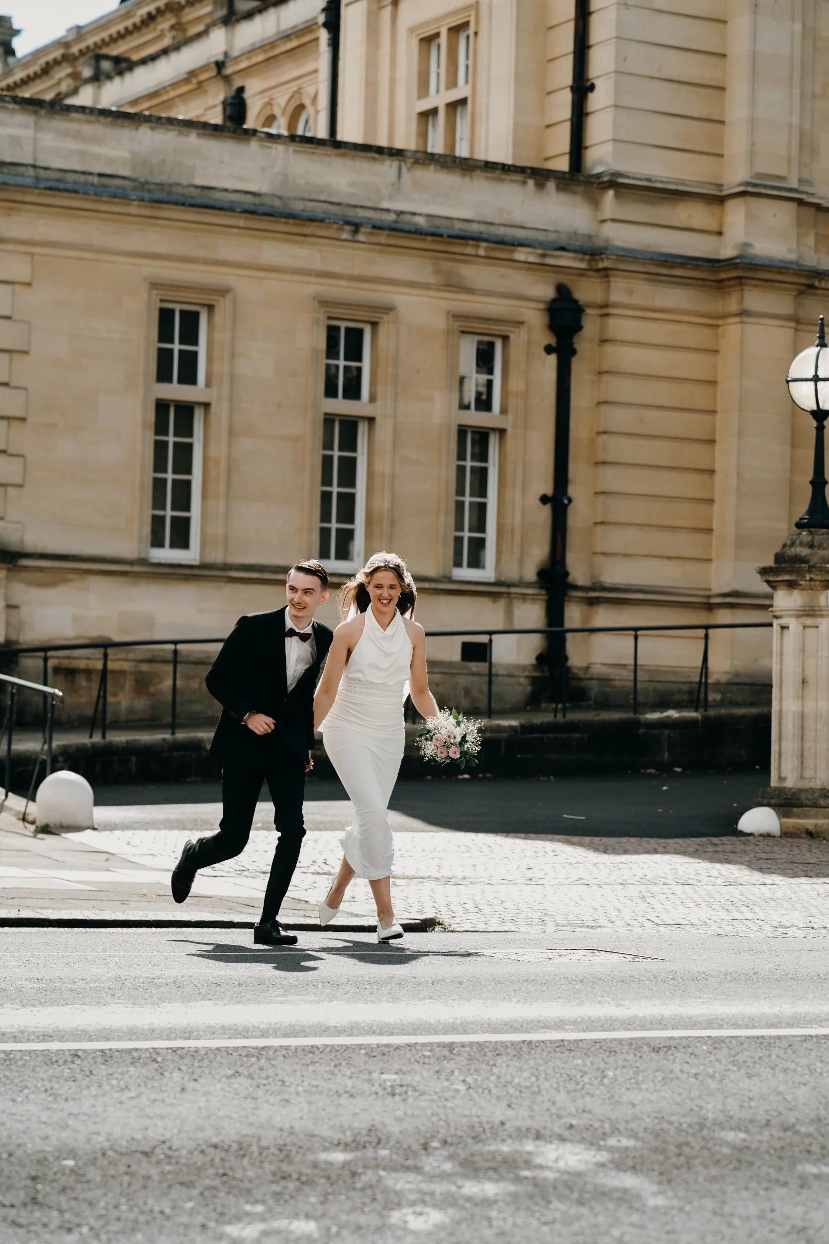 A newlywed couple is walking and laughing on a city street in front of a historic building, with the groom in a black tuxedo and the bride in a white gown holding a bouquet. Iris willow photography bar 131 Cheltenham 