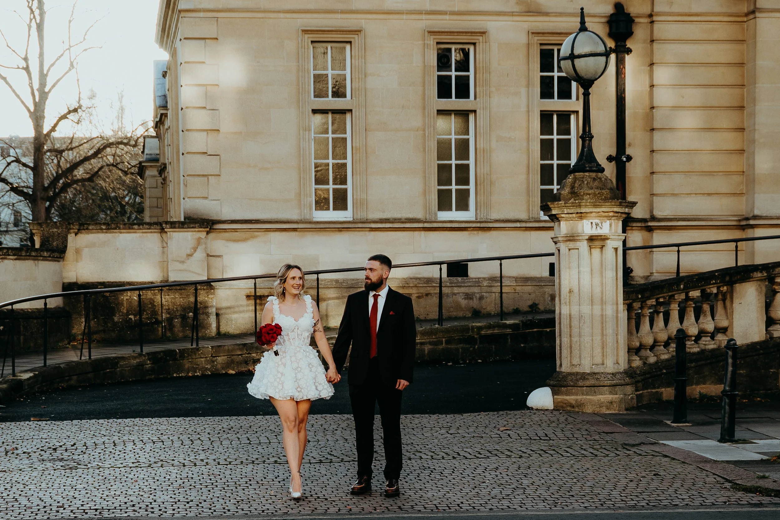 A bride and groom walking hand in hand outdoors near a historic building with large windows and a street lamp. The bride is holding a bouquet of red roses and wearing a lace wedding dress, while the groom is in a dark suit with a red tie.