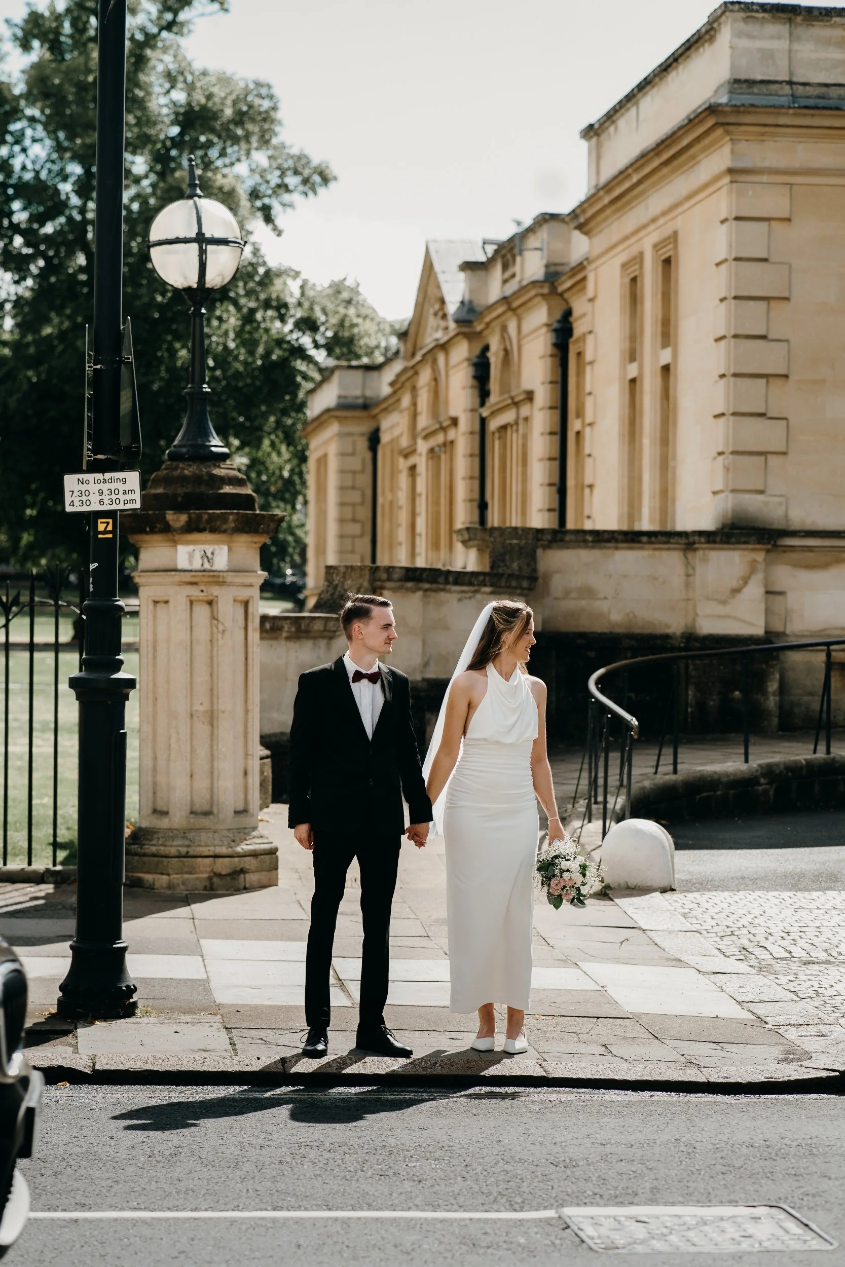 A bride and groom walking hand in hand on a city sidewalk, with a historic building and a street lamp in the background on a sunny day. Iris willow photography bar 131 Cheltenham 