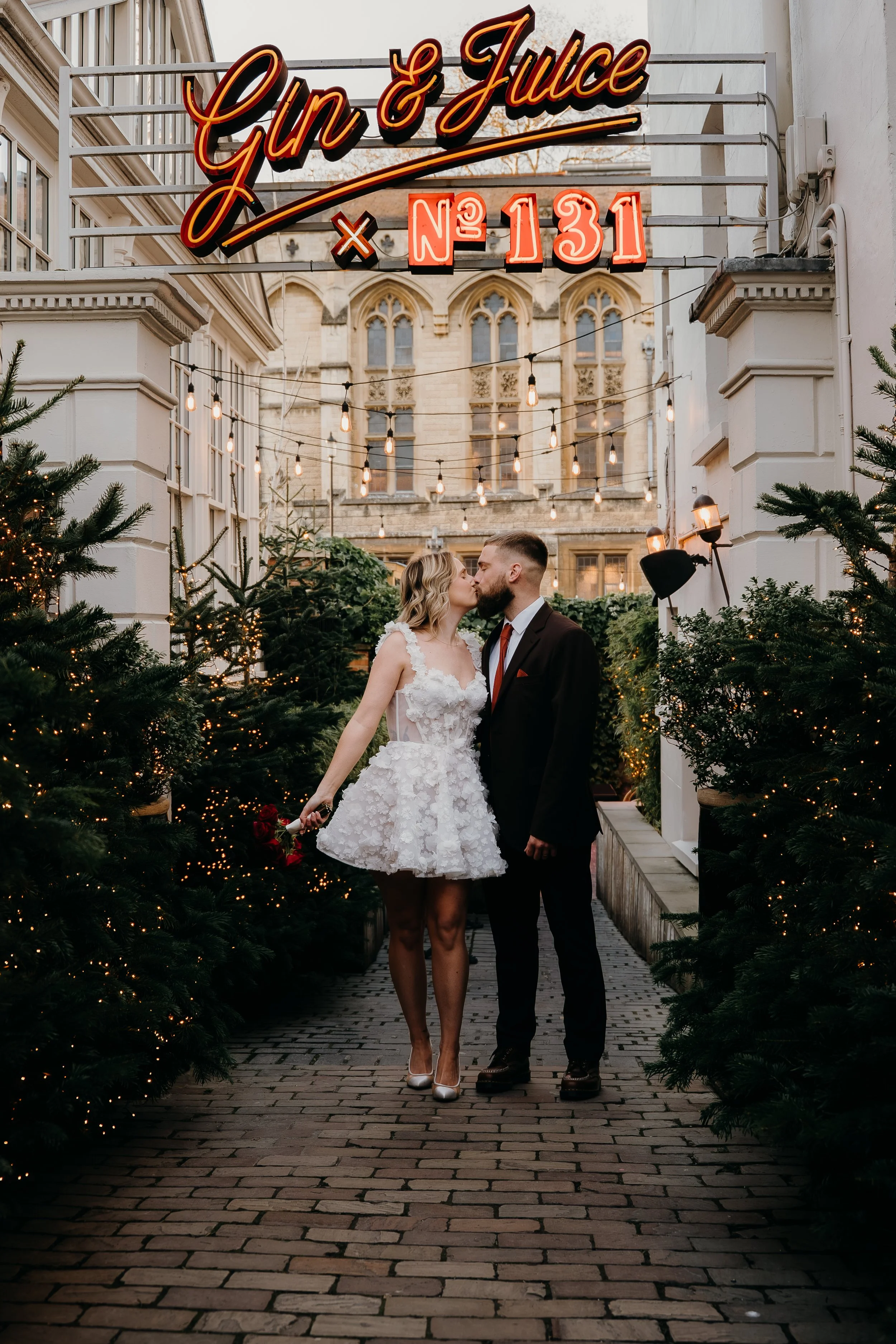 A couple, the woman in a white, lace wedding dress and the man in a dark suit, share a kiss as they walk through a decorated outdoor Christmas setting with string lights and Christmas trees. In the background, there is a historic building with gothic