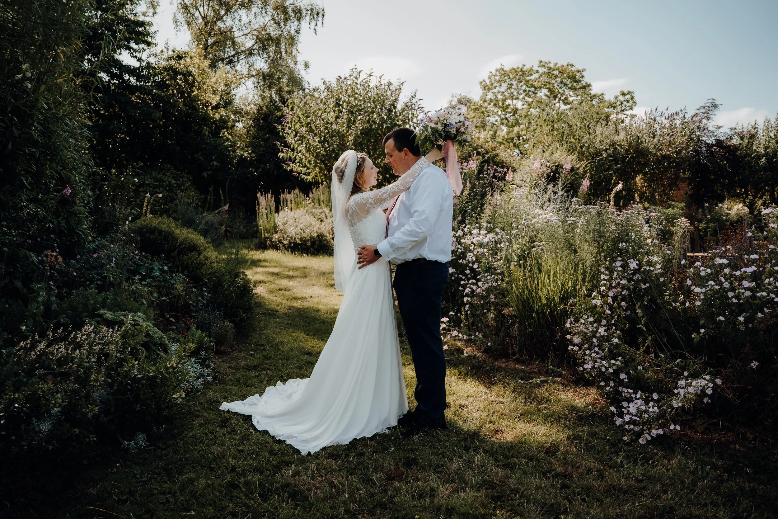 A bride and groom standing close together in a garden, facing each other, surrounded by flowering bushes and trees, with the bride wearing a white wedding dress and veil, and the groom in a white shirt and dark pants.