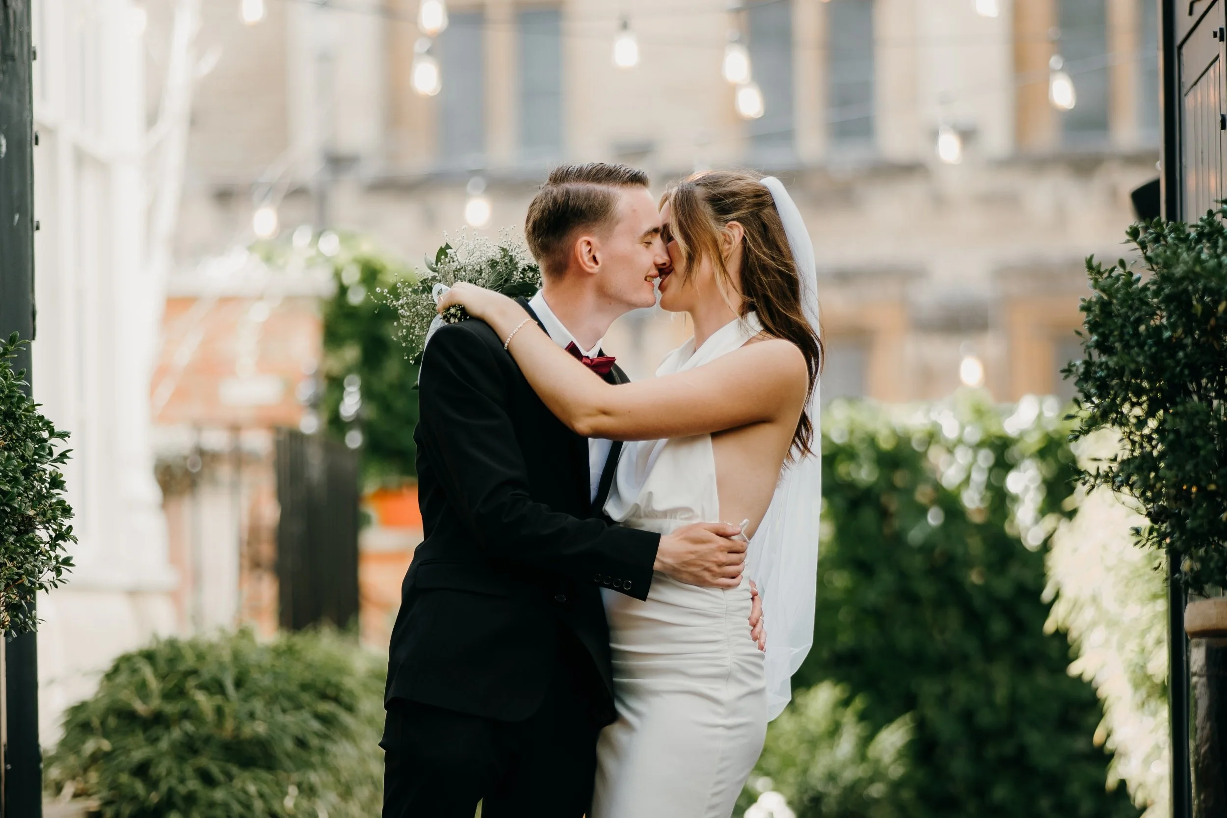 A bride and groom share a kiss outdoors, surrounded by greenery and string lights, during their wedding celebration. Iris willow photography bar 131 Cheltenham 
