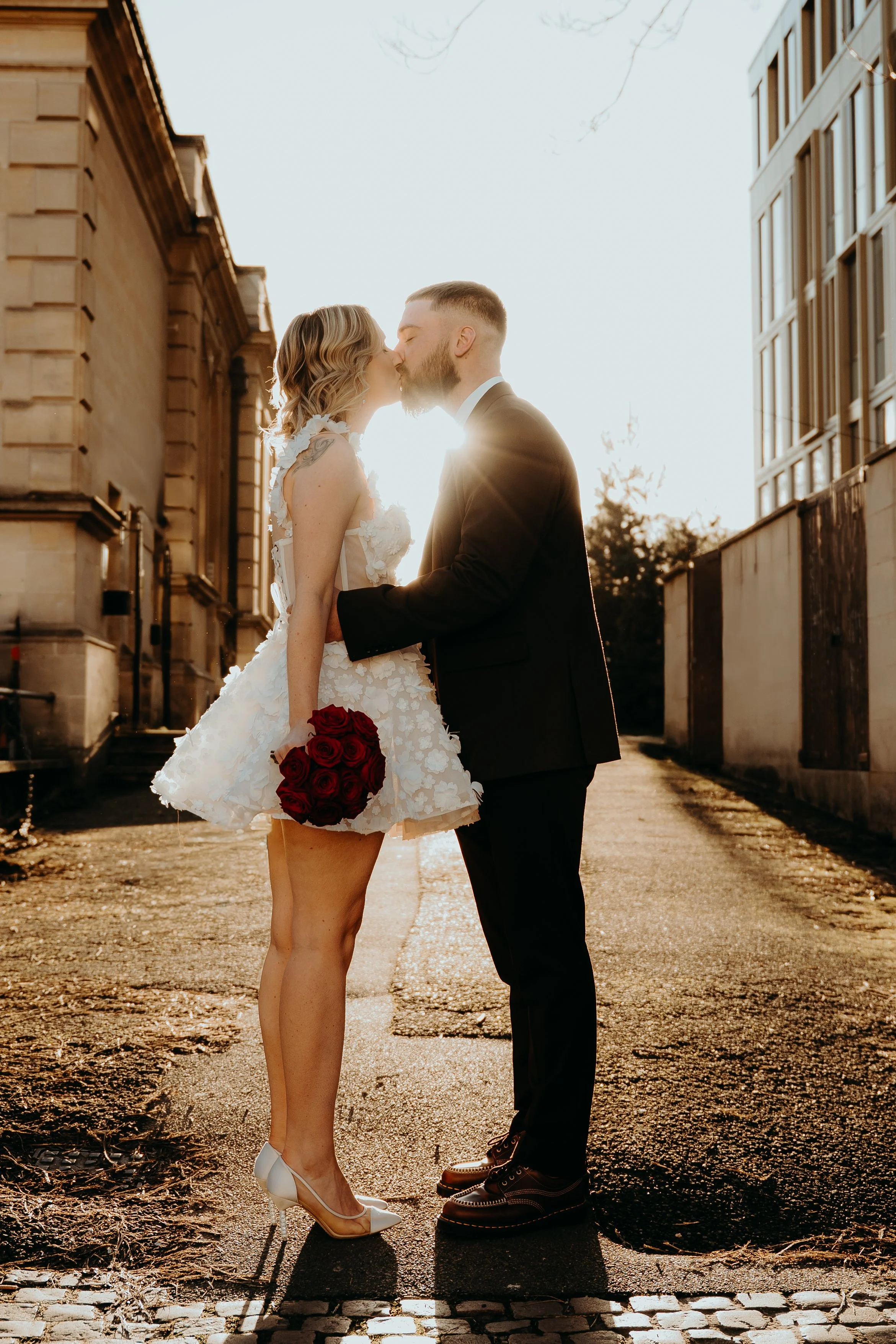A couple dressed in wedding attire sharing a kiss outdoors at sunset, with the bride holding a bouquet of red roses, between two buildings. Iris willow photography bar 131 Cheltenham