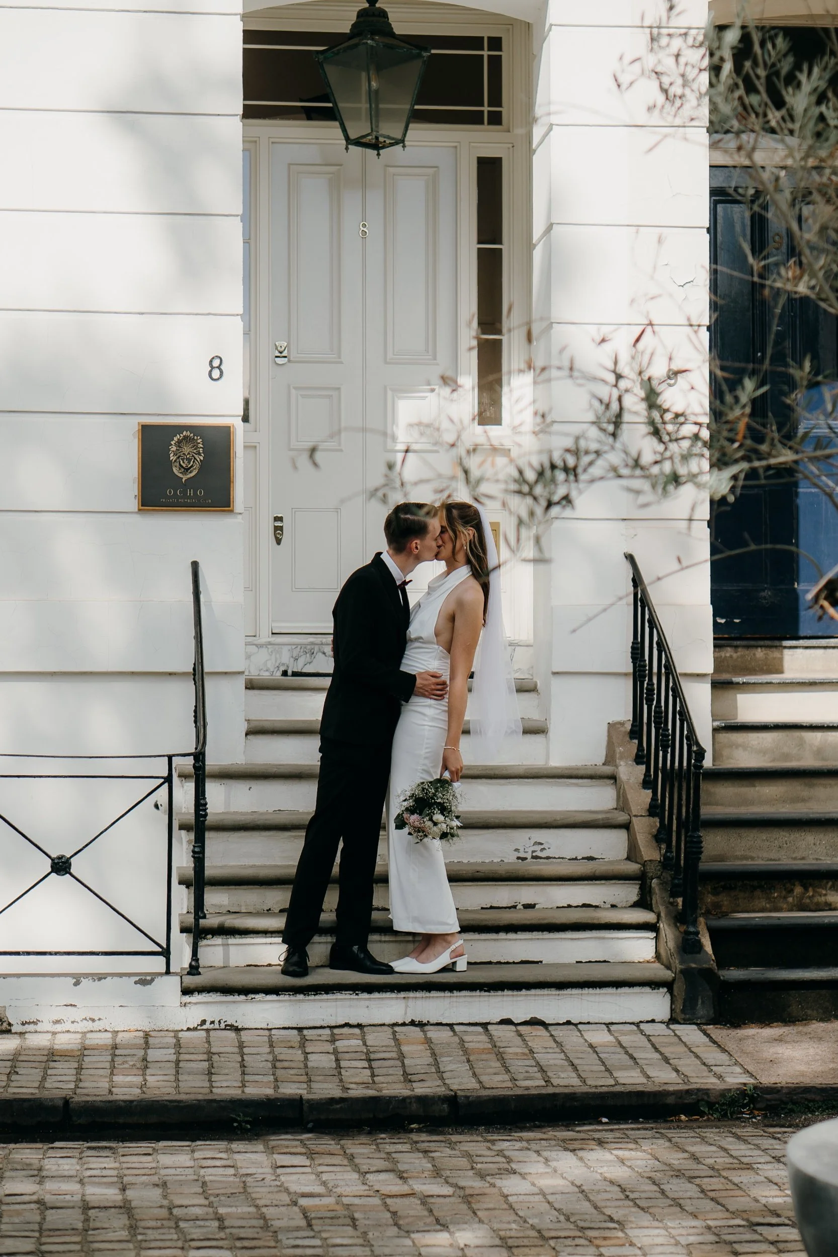 A newlywed couple, dressed in wedding attire, kisses on the steps of a white building with a black lantern hanging above the door. Iris willow photography bar 131 Cheltenham 