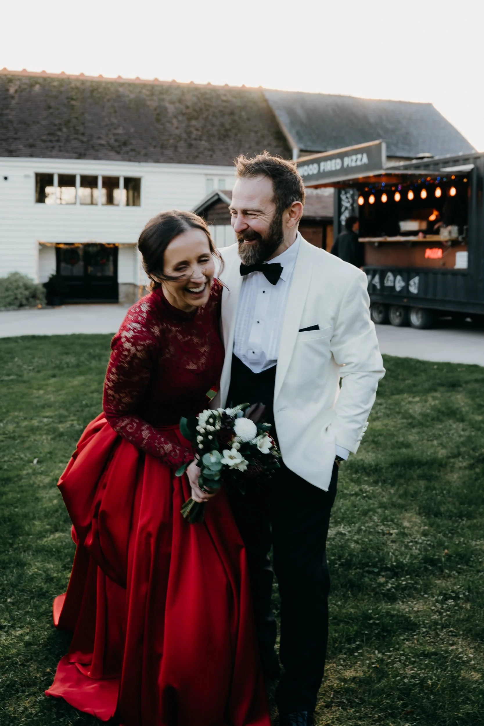 A man and woman in wedding attire laughing, standing outdoors on a grassy area near a food truck and white building during sunset.