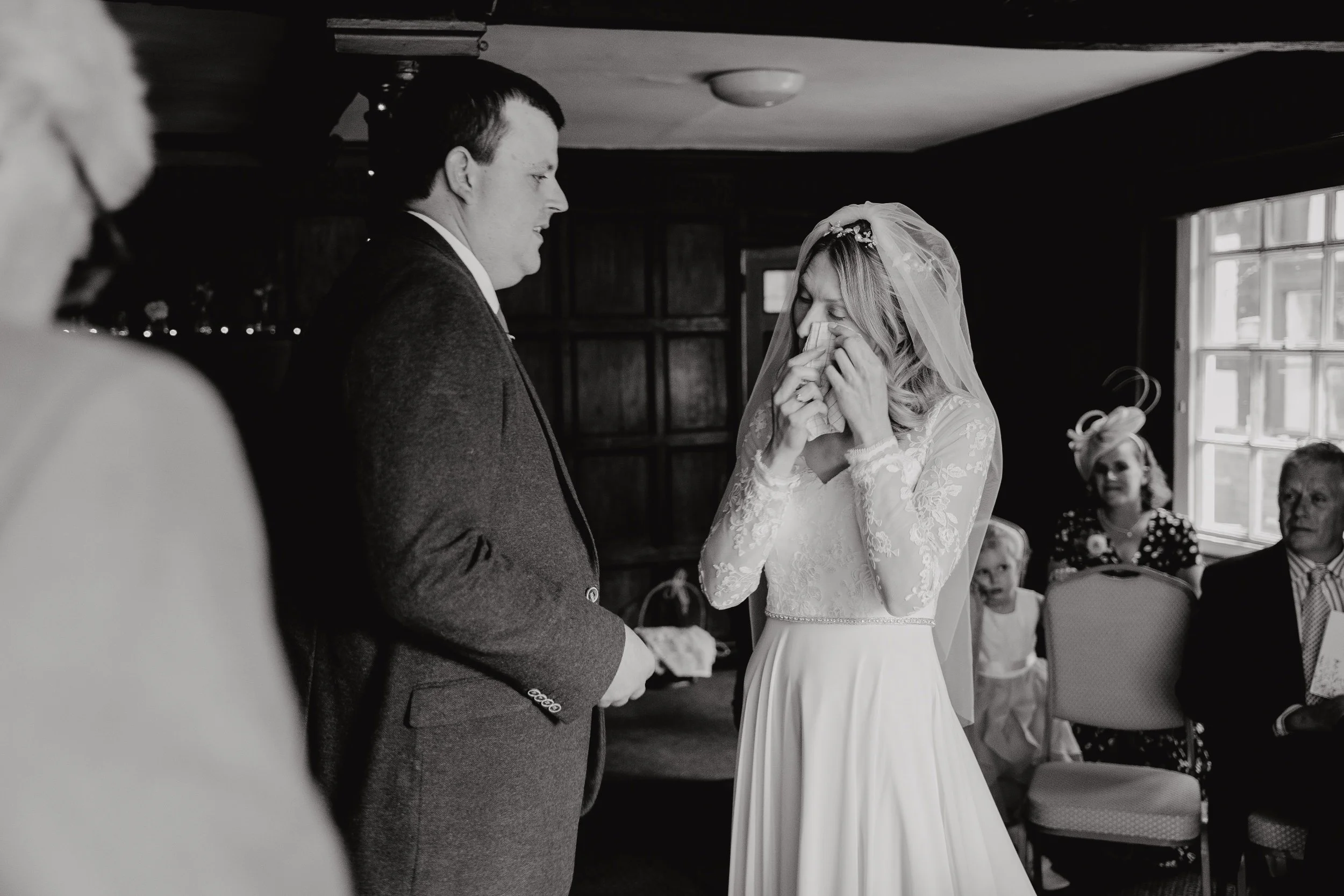 A bride wiping tears during her wedding ceremony, standing opposite a groom in a formal suit, indoors with guests visible in the background.