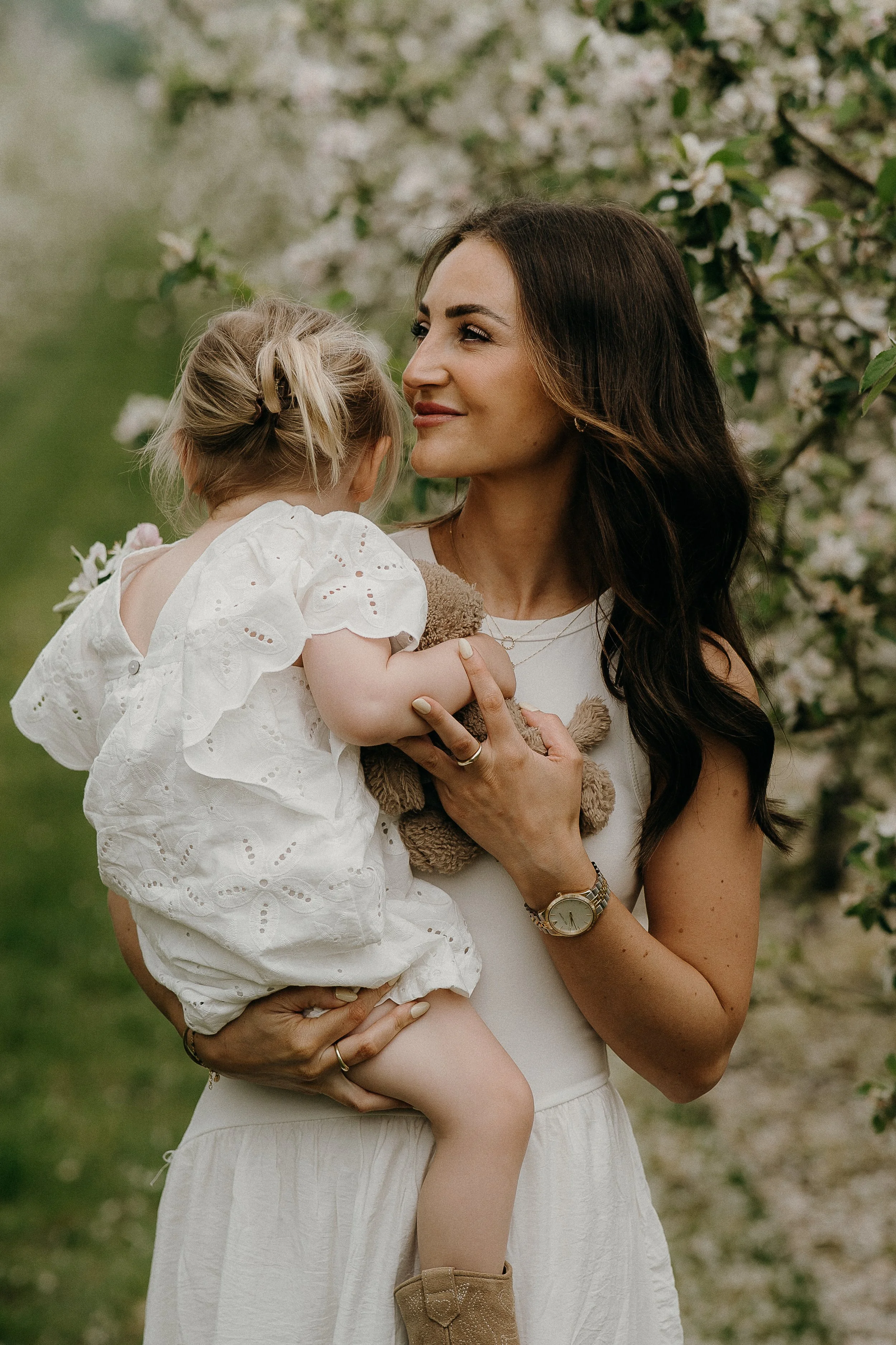 A woman with long dark hair holding a young blonde girl, both outdoors near blooming trees, sharing a moment of affection.