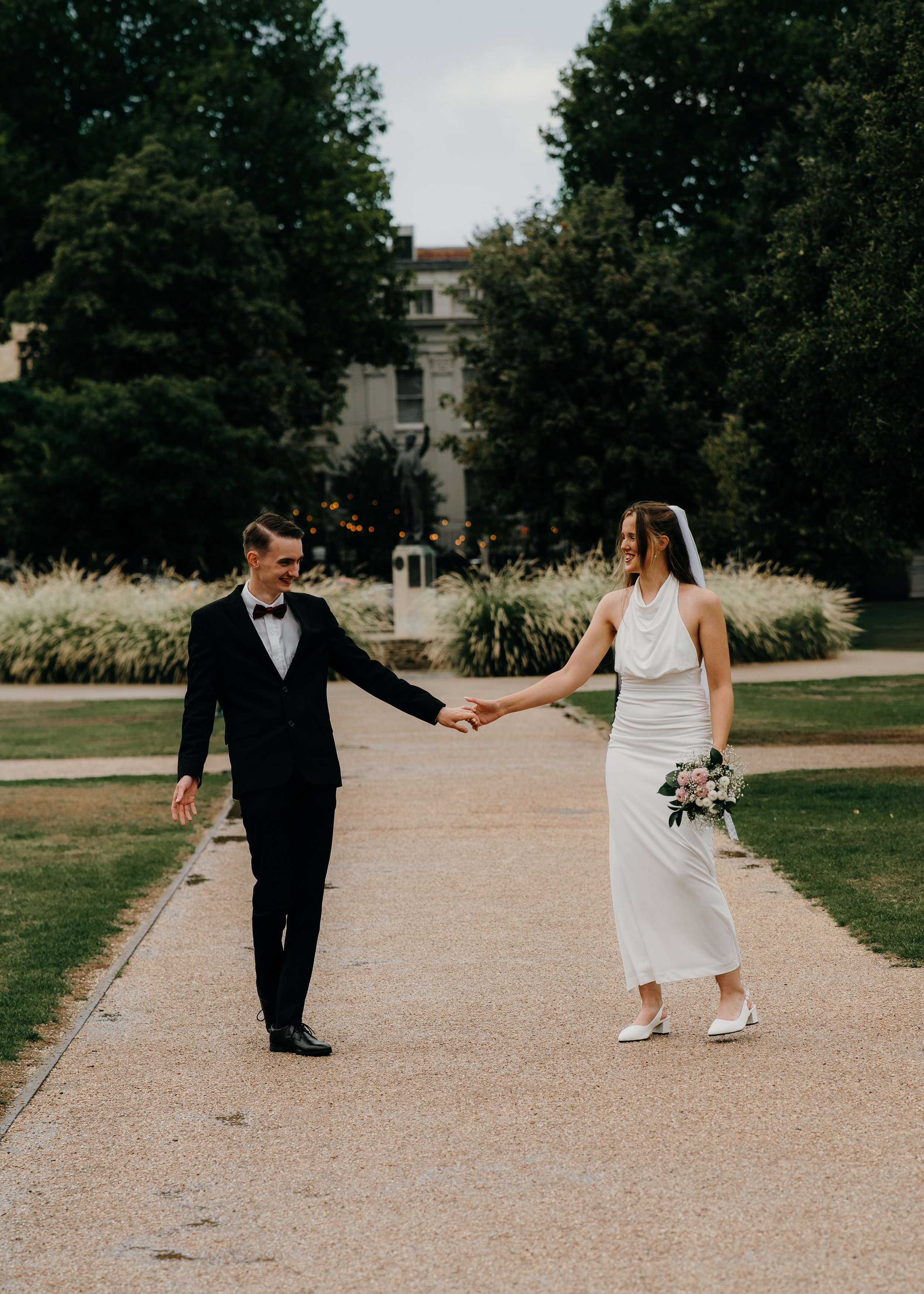 A man and woman in wedding attire holding hands and smiling in a park with trees and an old building in the background.