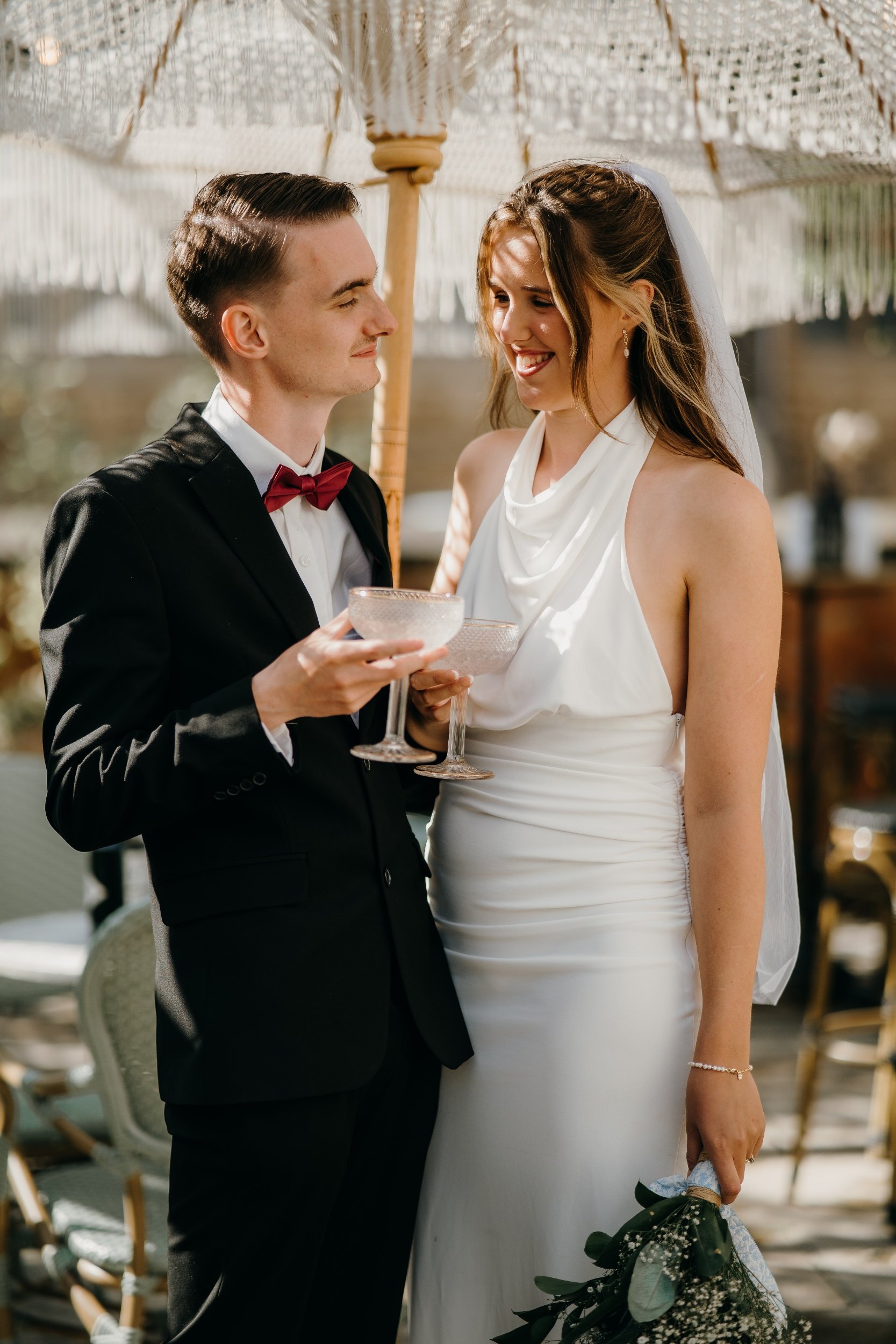 A couple at a wedding, the groom in a black suit with a red bow tie and the bride in a white dress holding a bouquet of flowers, smiling at each other under a paper parasol. Iris willow photography bar 131 Cheltenham 