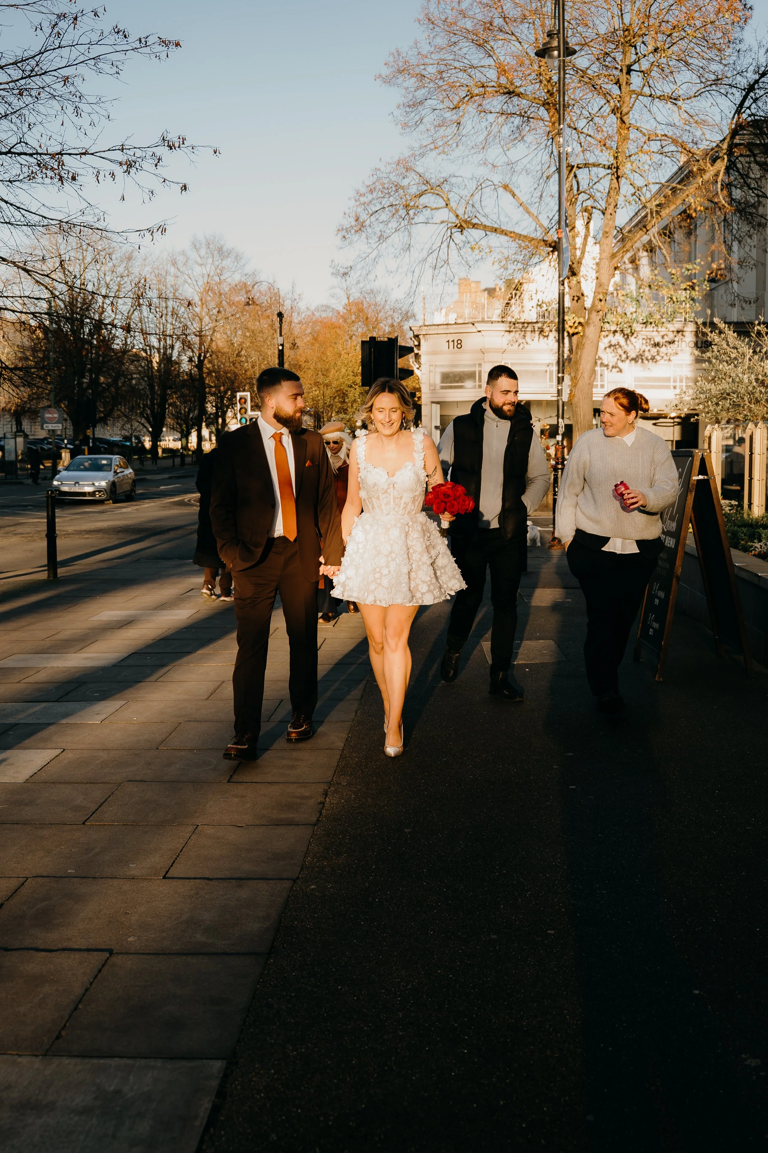 A woman in a white dress holding a red bouquet walking with three men on a city sidewalk during sunset, with leafless trees and buildings in the background.
