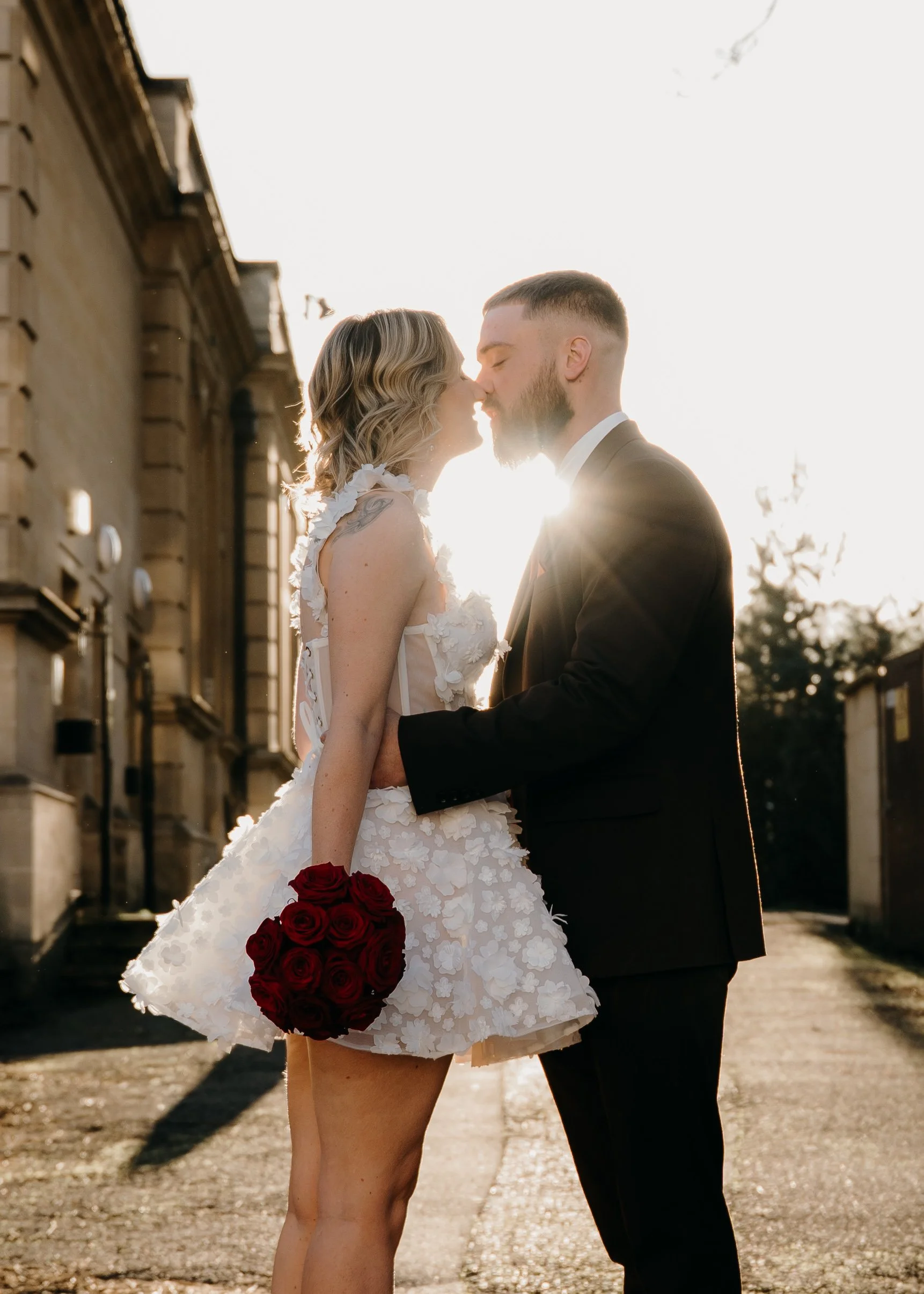 A couple dressed in wedding attire sharing a kiss outdoors at sunset, with the bride holding a bouquet of red roses.