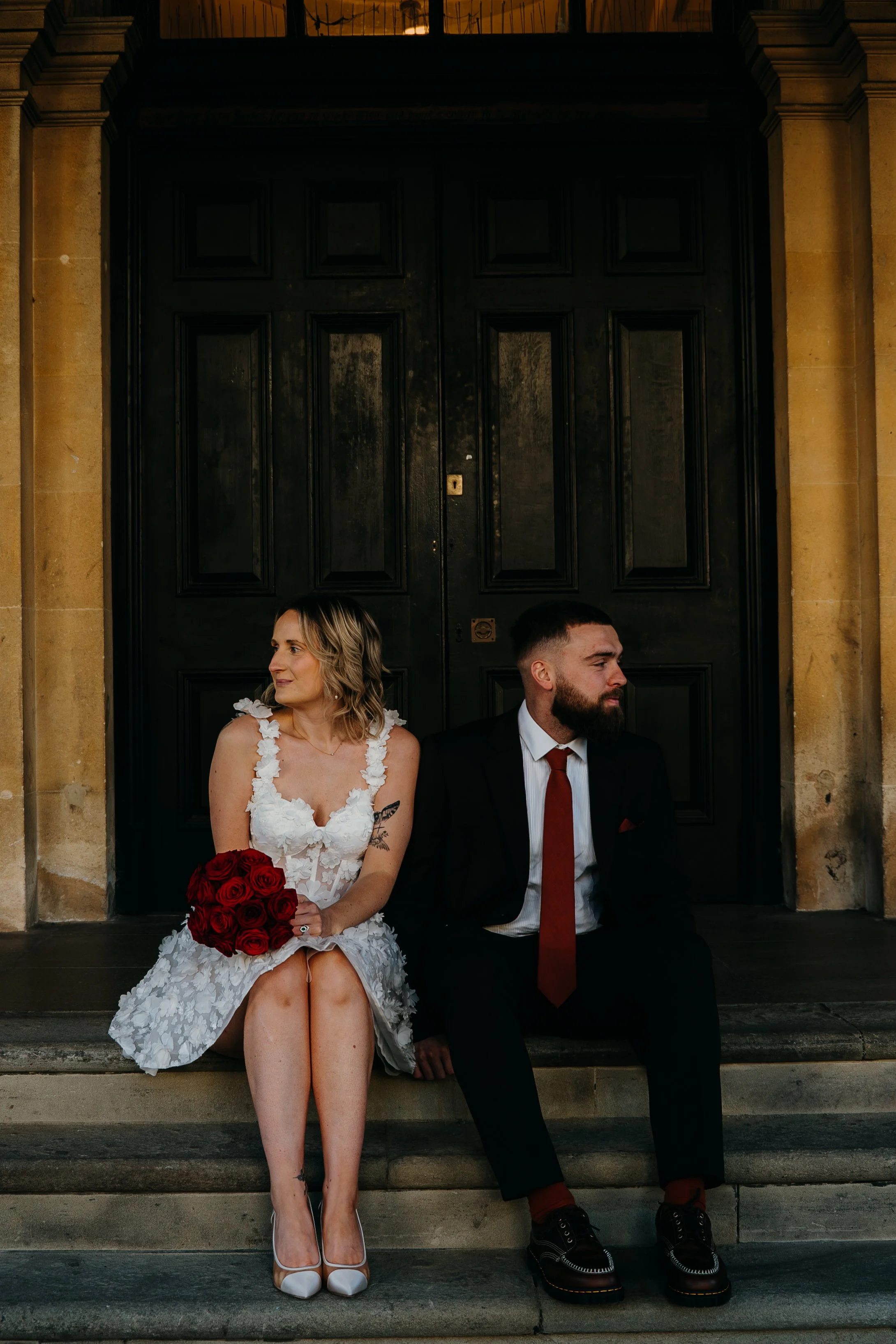 A woman in a white wedding dress holding a bouquet of red roses is sitting on steps in front of a large black door. Next to her, a man in a black suit with a red tie sits on the steps, looking to the side.