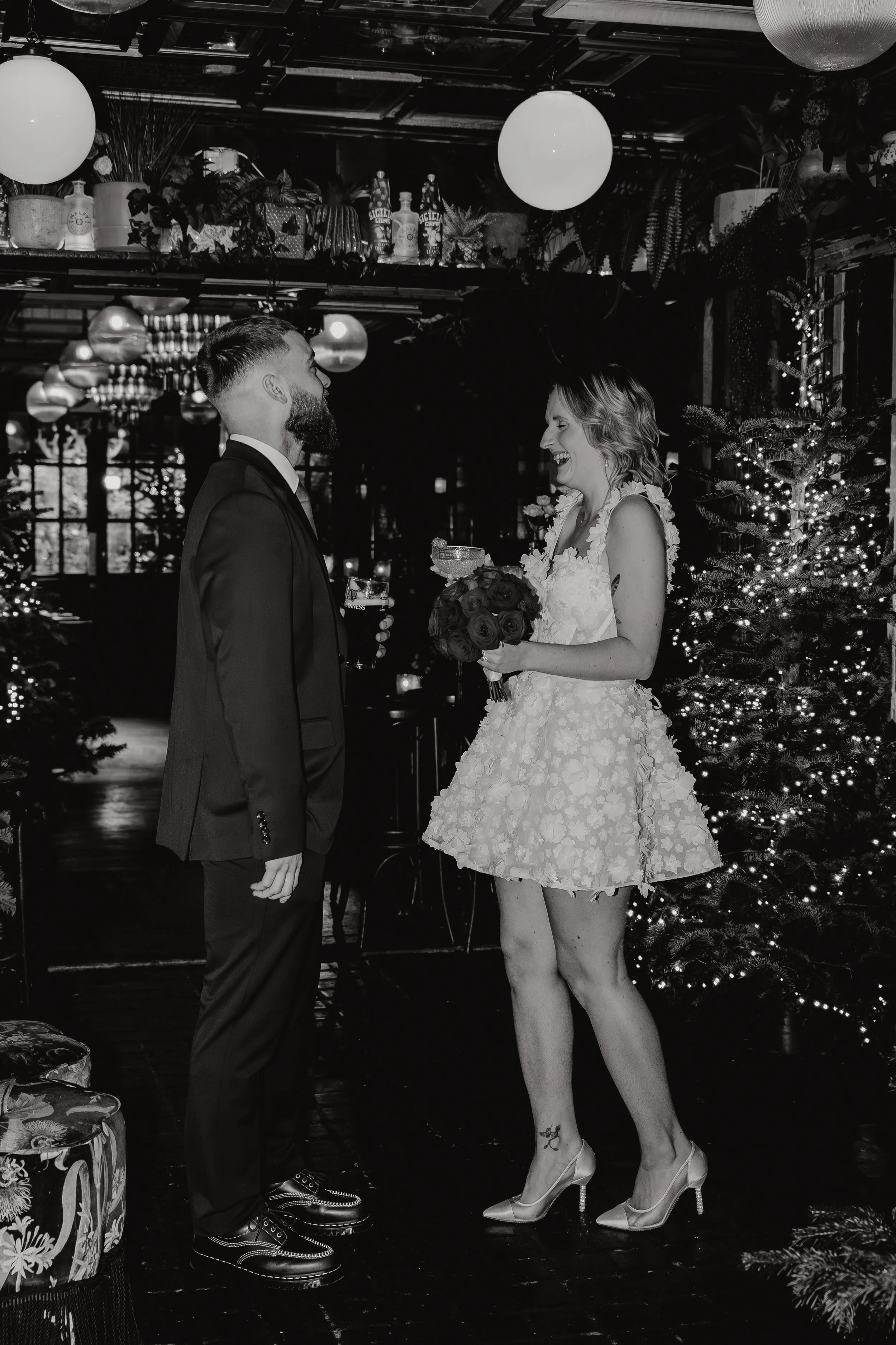 A man and woman stand facing each other in a festive setting decorated with Christmas trees, string lights, and hanging globe lights. The woman is wearing a short, floral-patterned dress and heels, holding a bouquet, and smiling. The man is dressed i