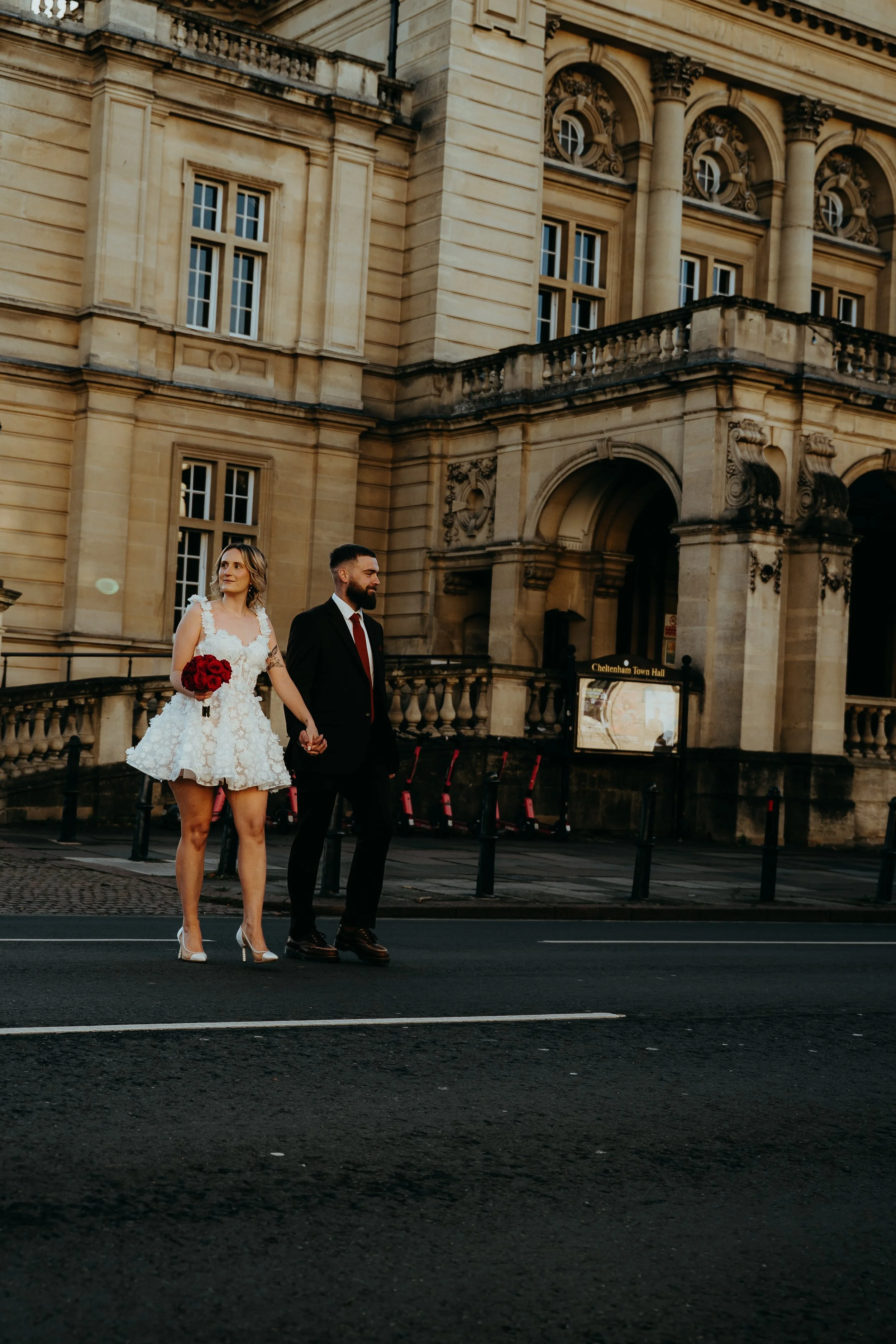 A couple walking hand in hand on a city street. The woman is wearing a white, floral dress with high heels, and holding a bouquet of red roses. The man is in a black suit with a white shirt and a red tie. They are in front of a historic, ornate stone