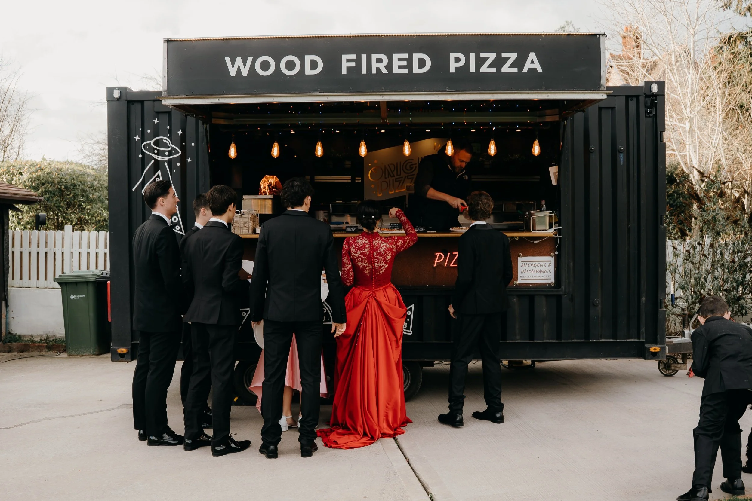 A black food truck with a sign that says 'Wood Fired Pizza' serves customers. Several people in formal attire are lined up to order, with one woman in a red dress and others in black suits waiting.