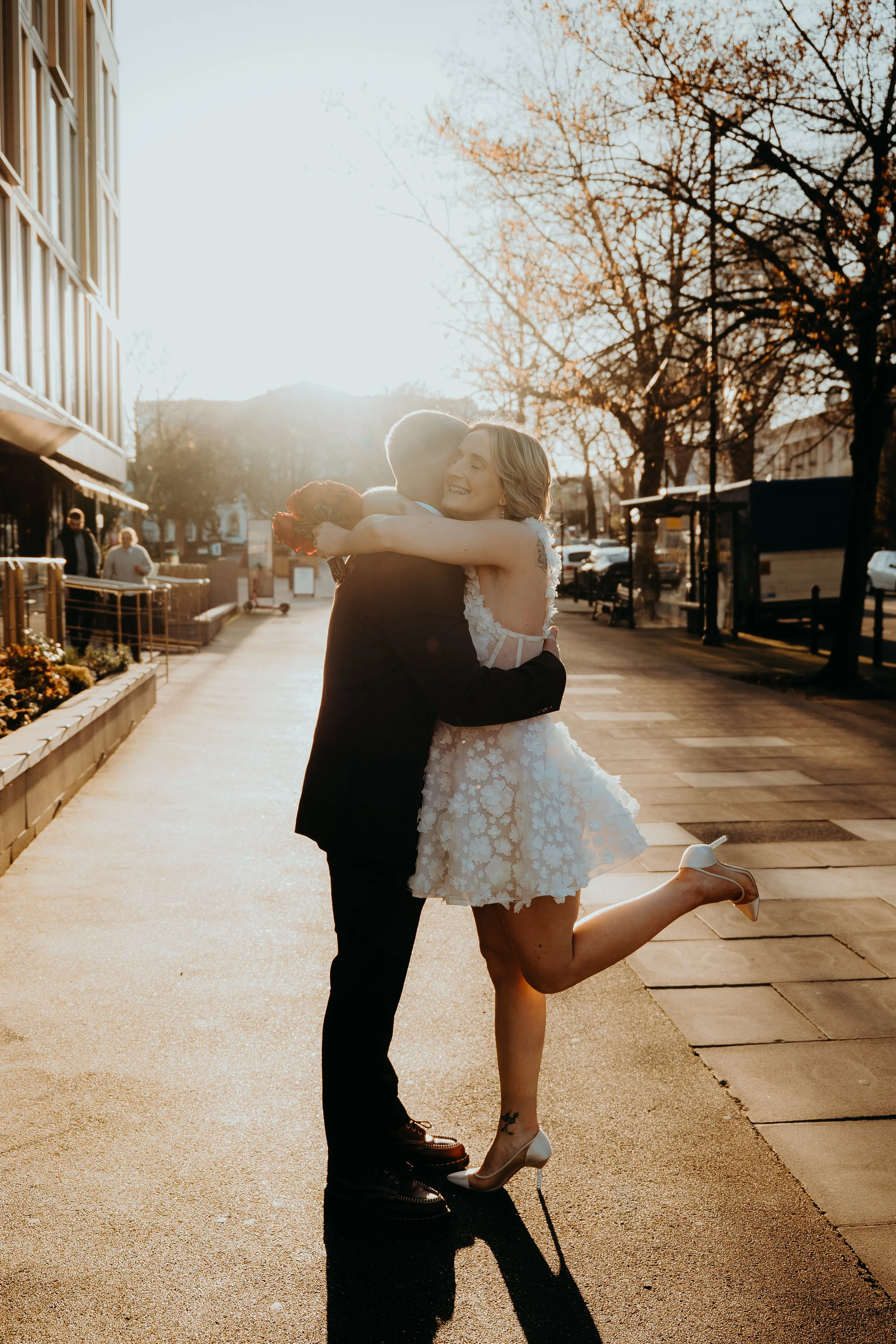 A couple celebrates their wedding outdoors during sunset. The woman, in a white lace dress and heels, is kneeling on one leg with a joyful expression. The man, in a dark suit, is hugging her. Trees with bare branches are visible in the background, along with a few pedestrians and parked cars.