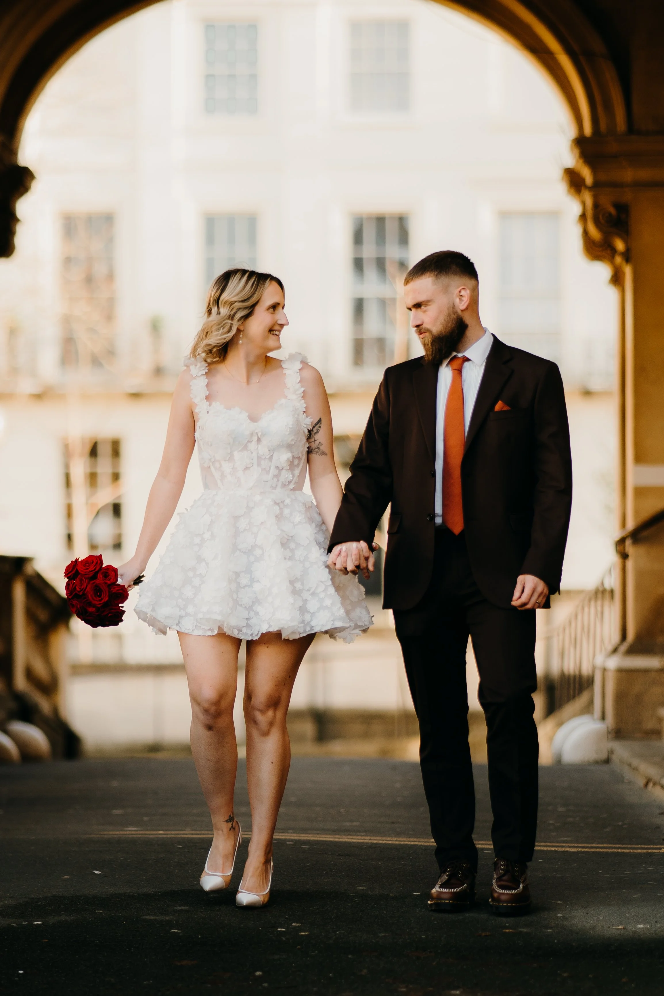 A couple walking hand in hand through an archway. The woman is wearing a white wedding dress with floral details and holding a bouquet of red roses. The man is dressed in a black suit with a white shirt and a red tie, and has a beard. They are smilin
