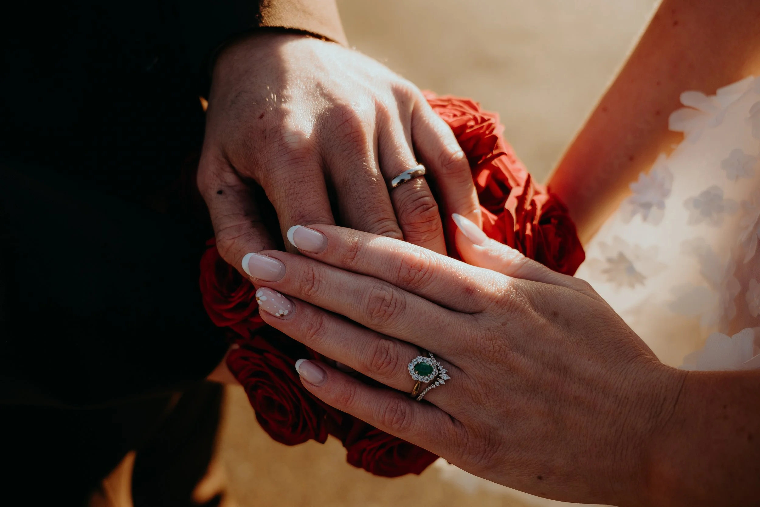 Close-up of two hands, one with a wedding ring, gently holding each other, with a red flower and a white flower in the background. Iris willow photography bar 131 Cheltenham