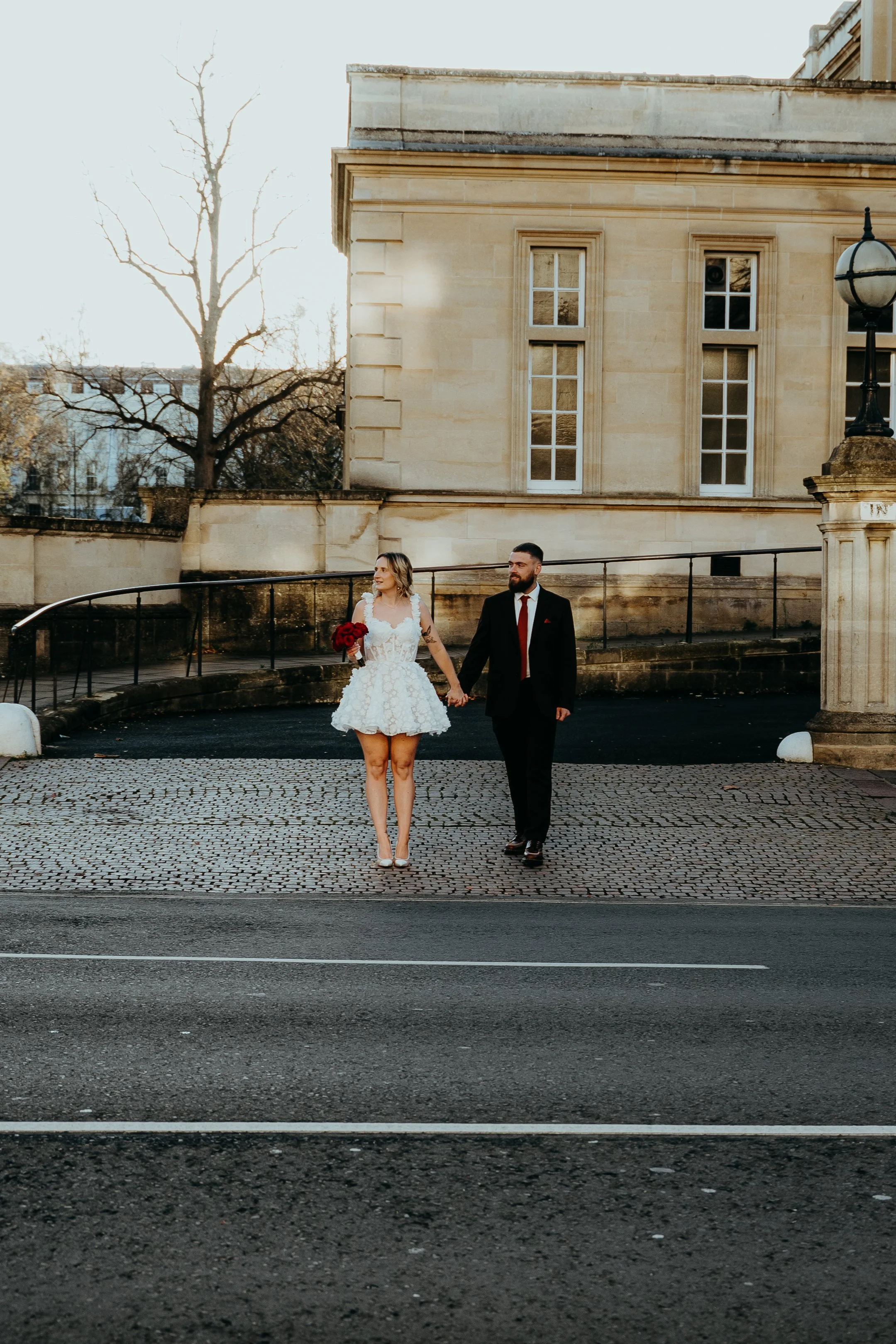 A couple in wedding attire walking hand in hand, holding a bouquet of red roses, in front of a historic building with large windows and a streetlamp. The scene is set outdoors in daylight.
