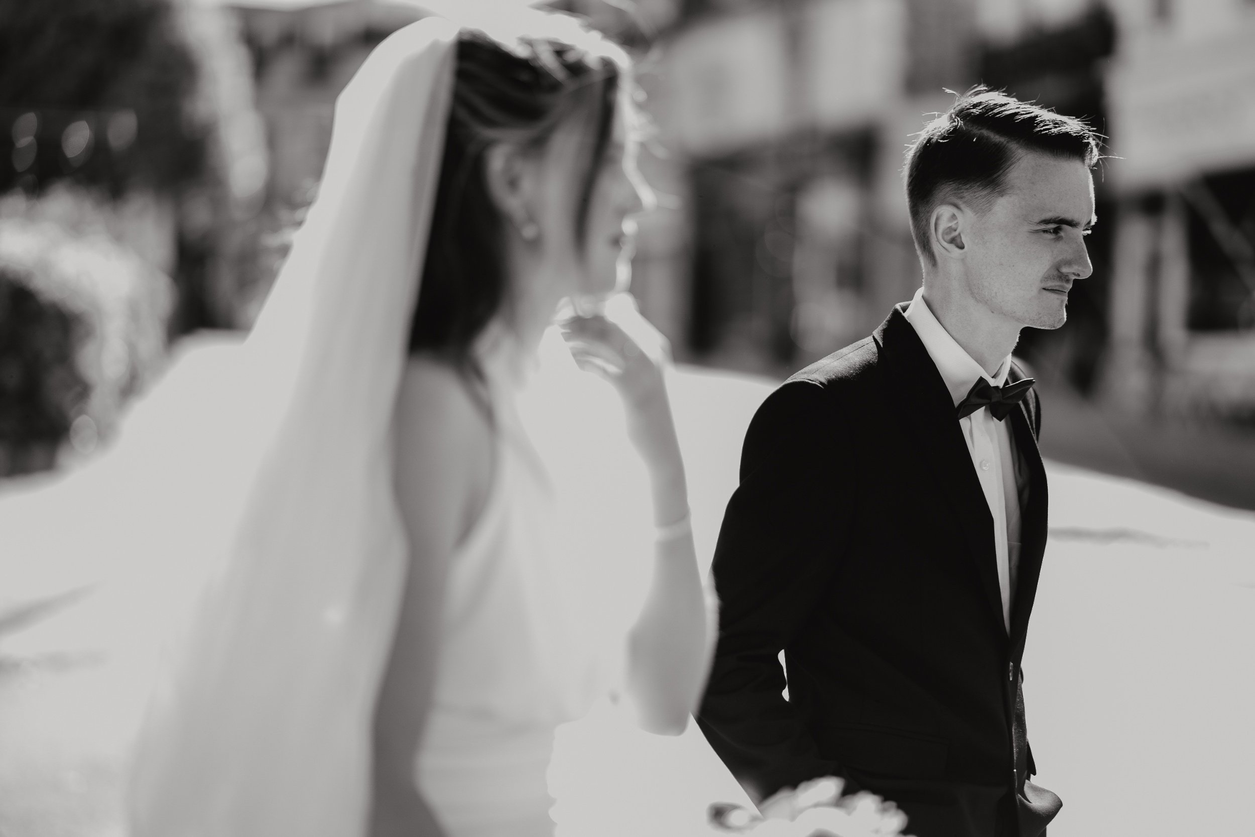 Black and white photo of a man in a tuxedo with a bow tie, standing outdoors with sunlight highlighting his face and hair. In the blurred foreground, a woman wearing a wedding dress. Iris willow photography bar 131 Cheltenham 