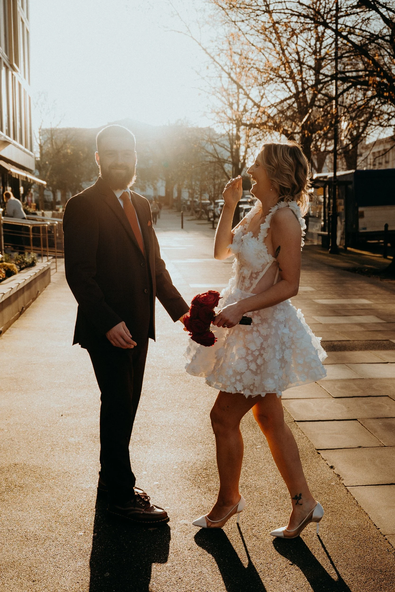 A couple dressed in wedding attire standing outdoors during sunset, holding hands and sharing a moment, with the woman laughing and holding a bouquet of red roses.