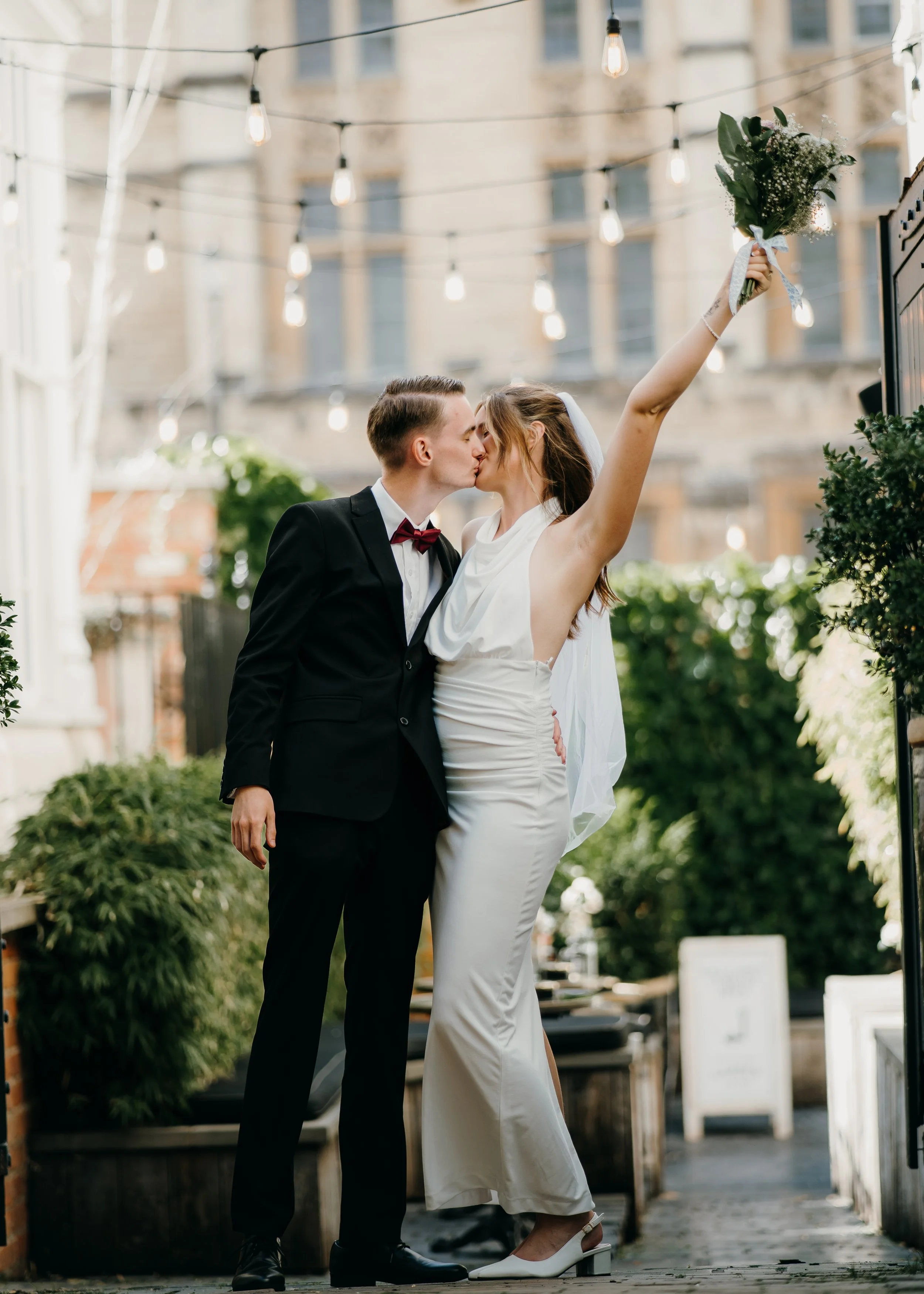 A couple wearing wedding attire, sharing a kiss outdoors beneath string lights, with the bride holding a bouquet aloft. The groom is in a black tuxedo and the bride in a white wedding dress and heels.