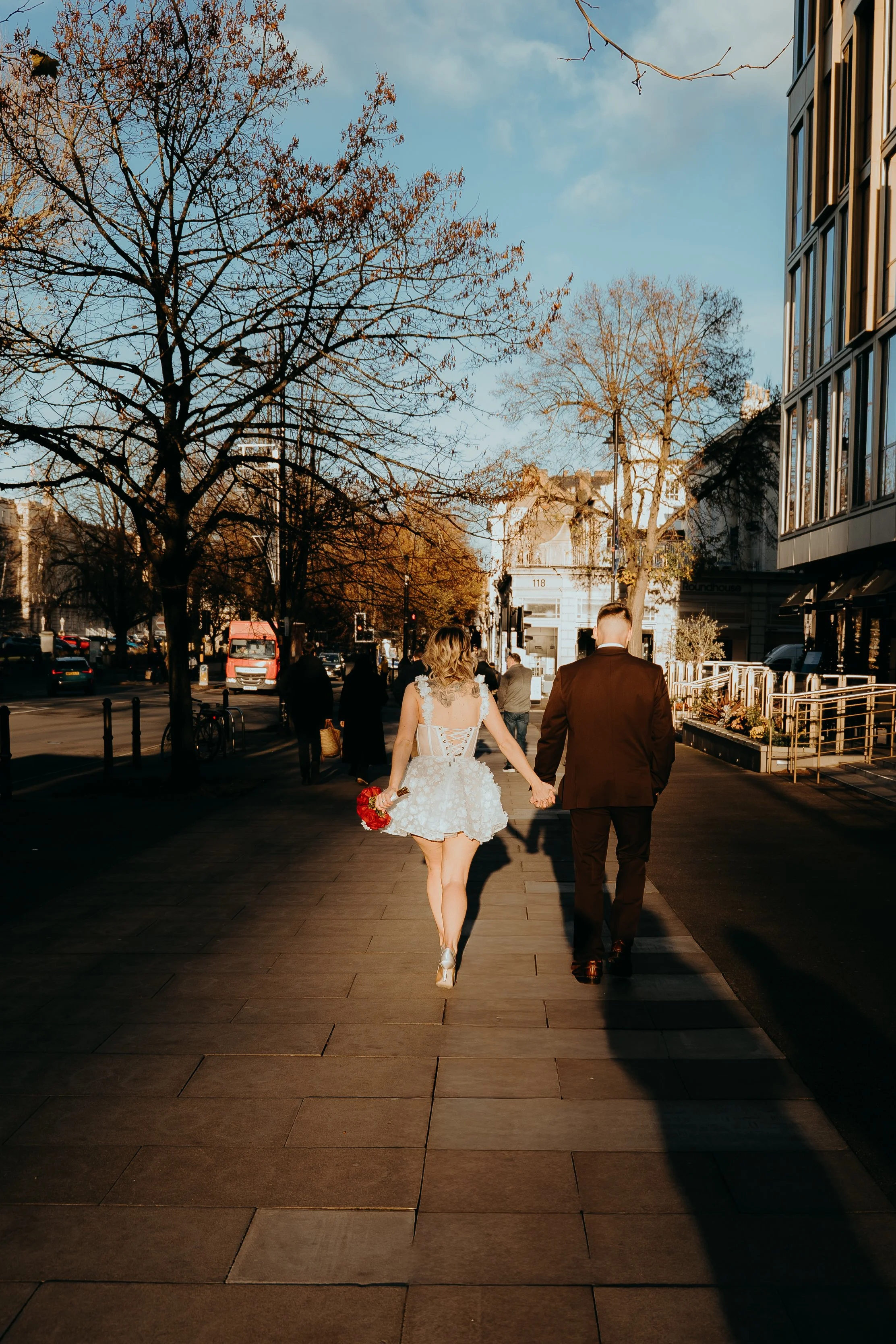 A bride and groom hand in hand on a city sidewalk with trees and buildings around, during late afternoon. Iris willow photography bar 131 Cheltenham