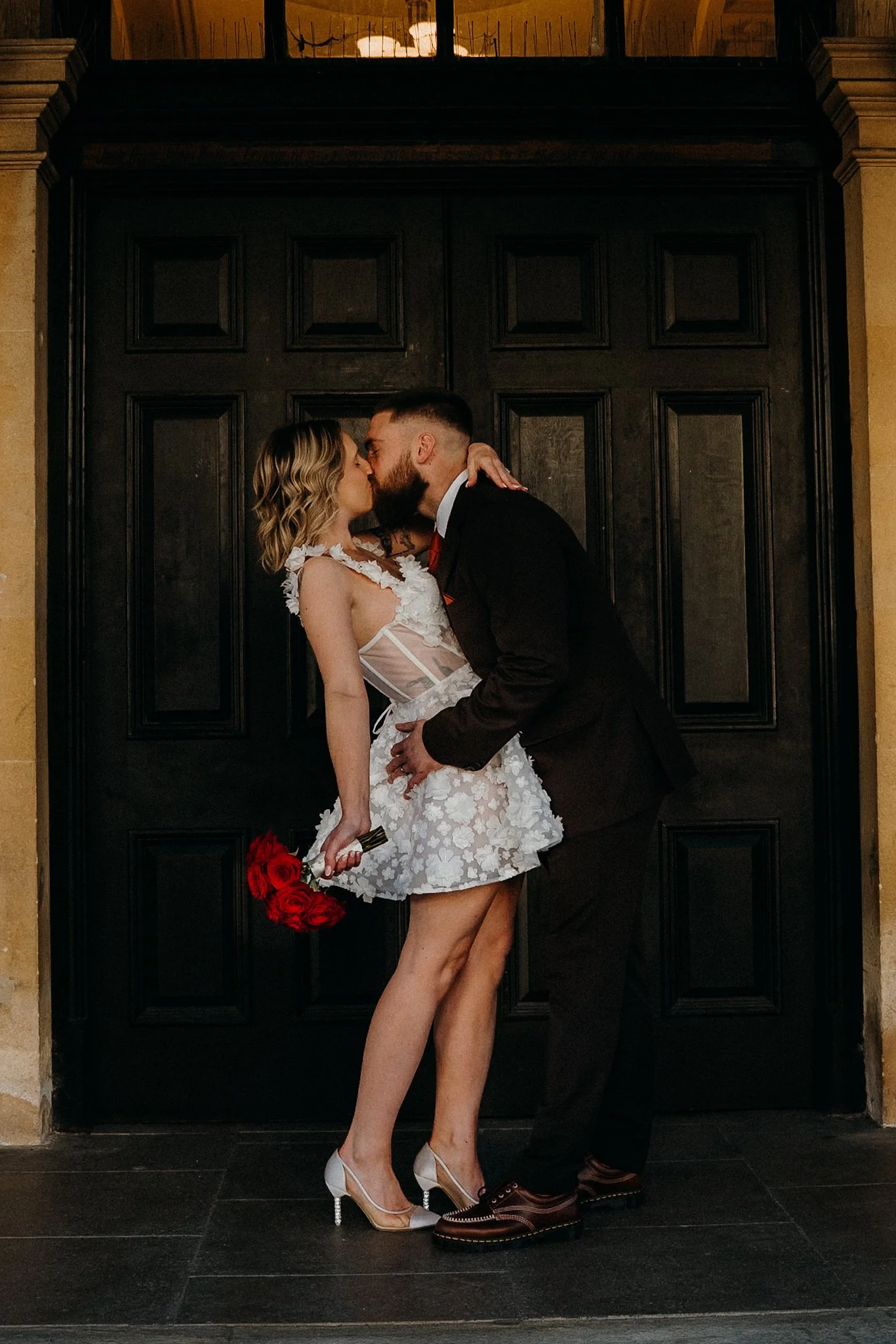 A couple is kissing in front of a dark wooden door. The woman is wearing a white lace dress and high heels, holding a bouquet of red roses. The man is dressed in a dark suit and shoes. The scene appears to be romantic, possibly a wedding or engagement photo.