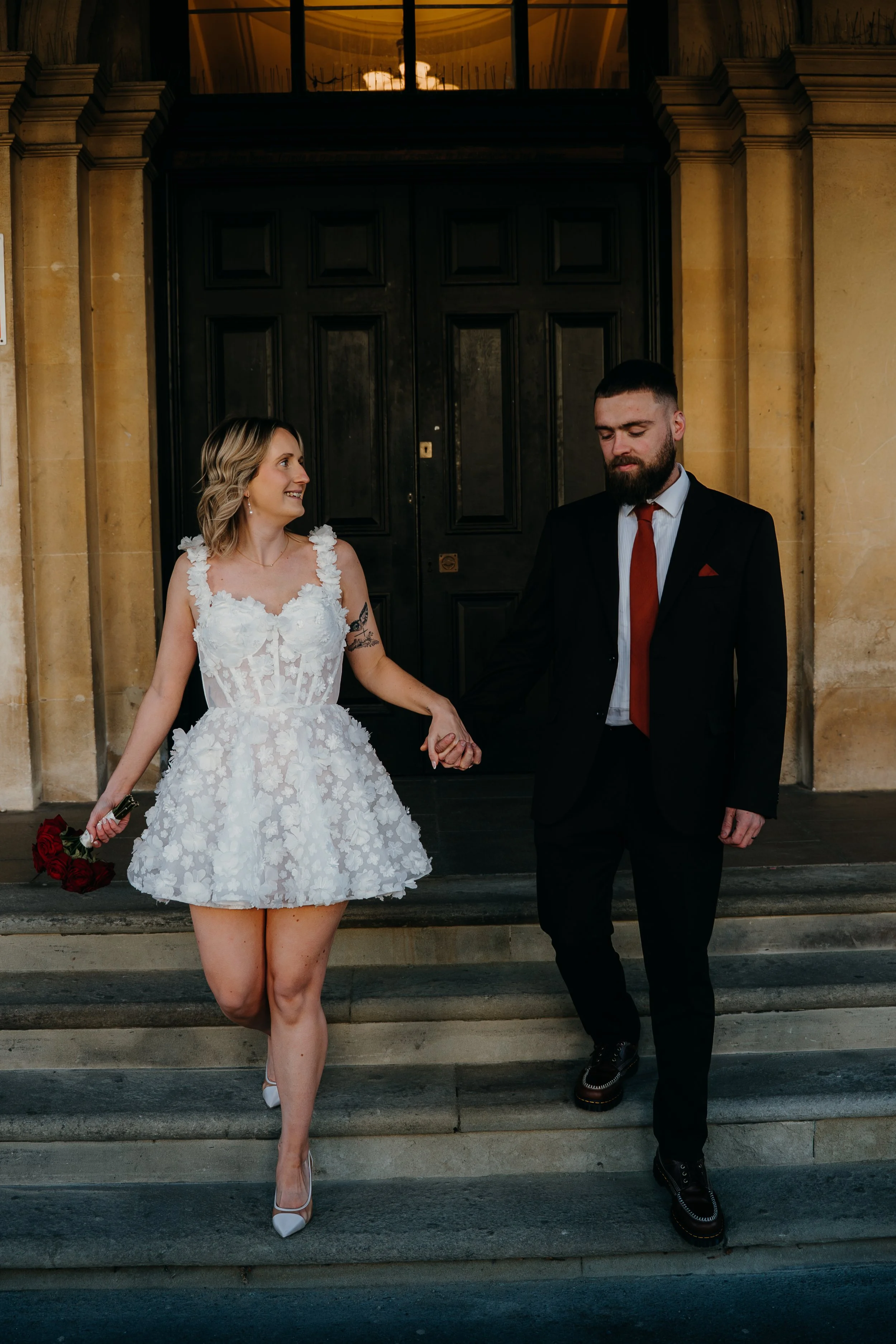 A bride and groom holding hands on the steps of a building at night. The bride is wearing a white floral dress, holding a bouquet of red roses, and white high heels. Iris willow photography bar 131 Cheltenham