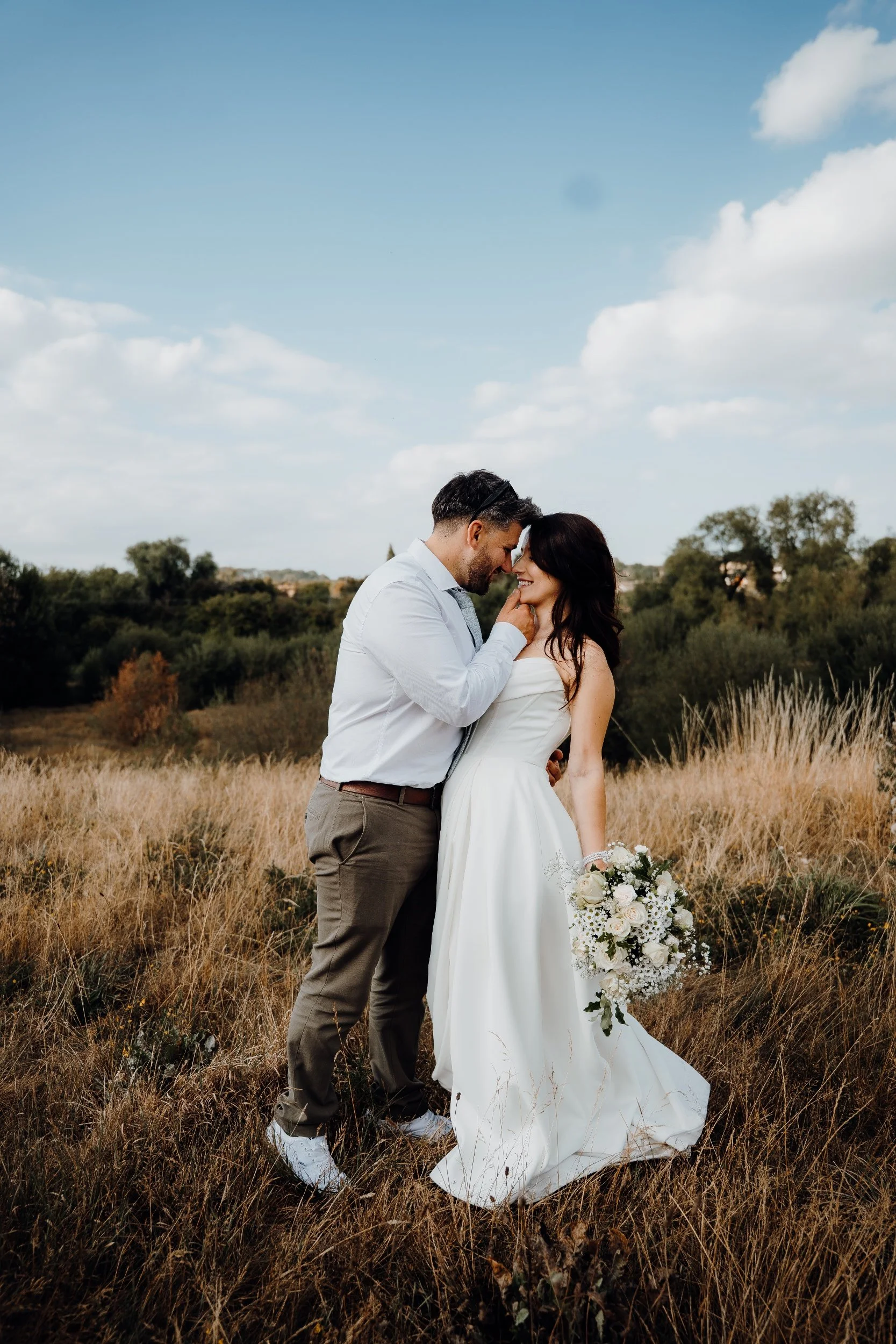 A wedding couple stands close together in a field, the man gently touching the woman's face, both smiling as they look into each other's eyes. The woman wears a white wedding dress and holds a bouquet of flowers, while the man wears a white shirt and gray trousers. The background features tall, golden grass, trees, and a partly cloudy sky.
