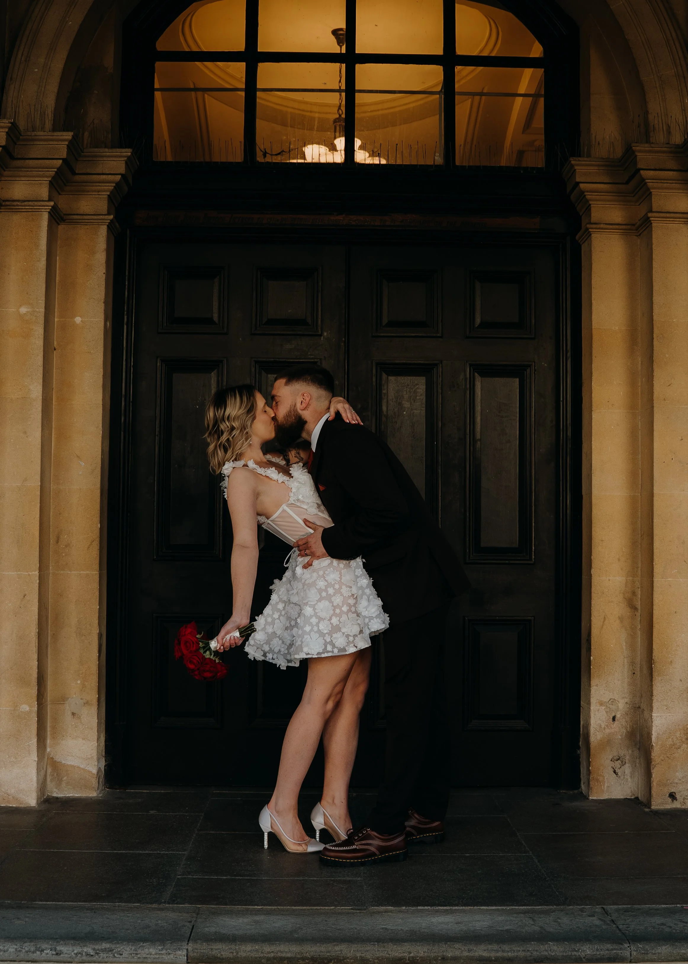 A couple in wedding attire sharing a kiss in front of a large black door with a window above.  Iris willow photography bar 131 Cheltenham