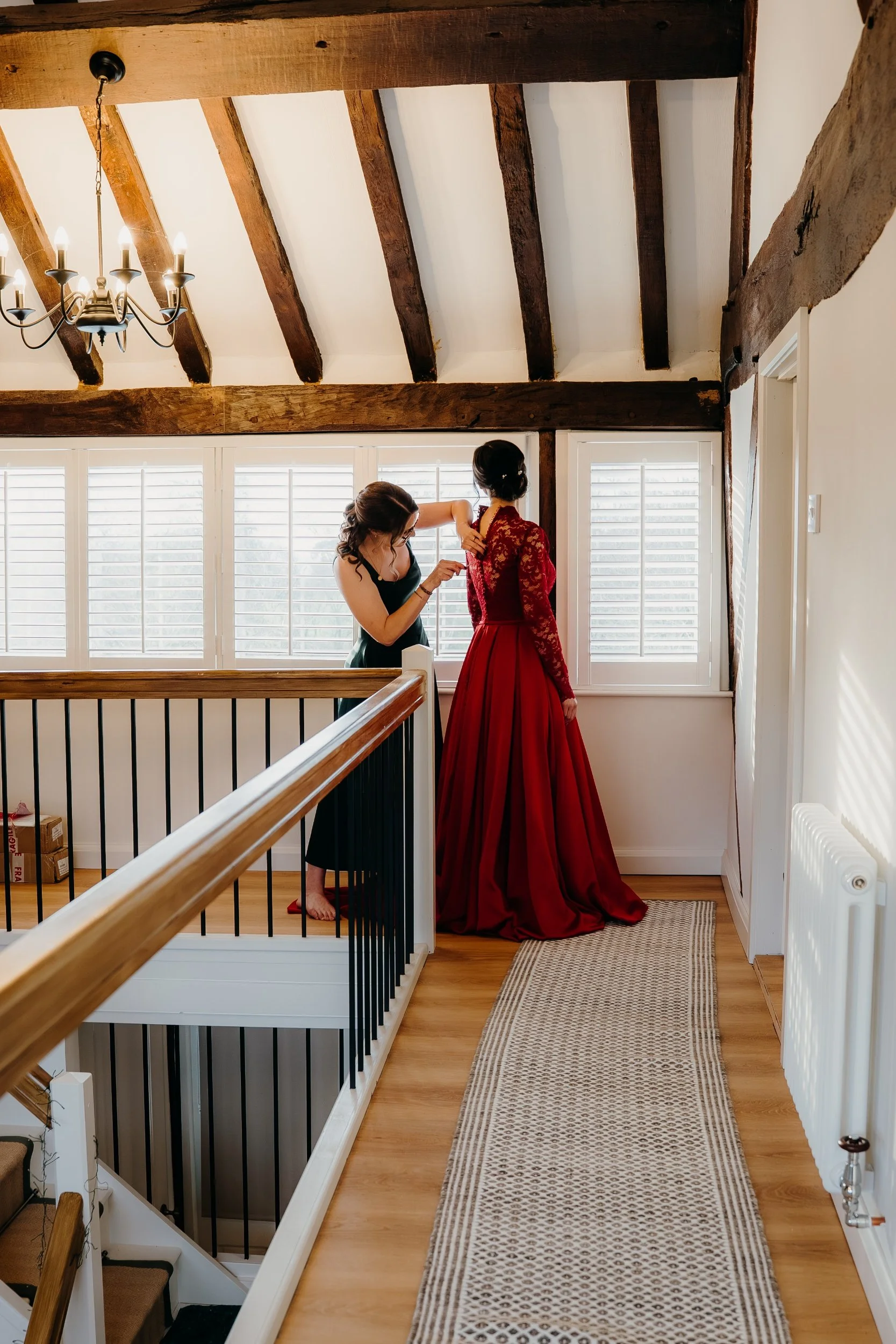 A woman in a black dress helping another woman in a red gown get ready by adjusting her dress near a window with white shutters, in a room with wooden beams and a chandelier.
