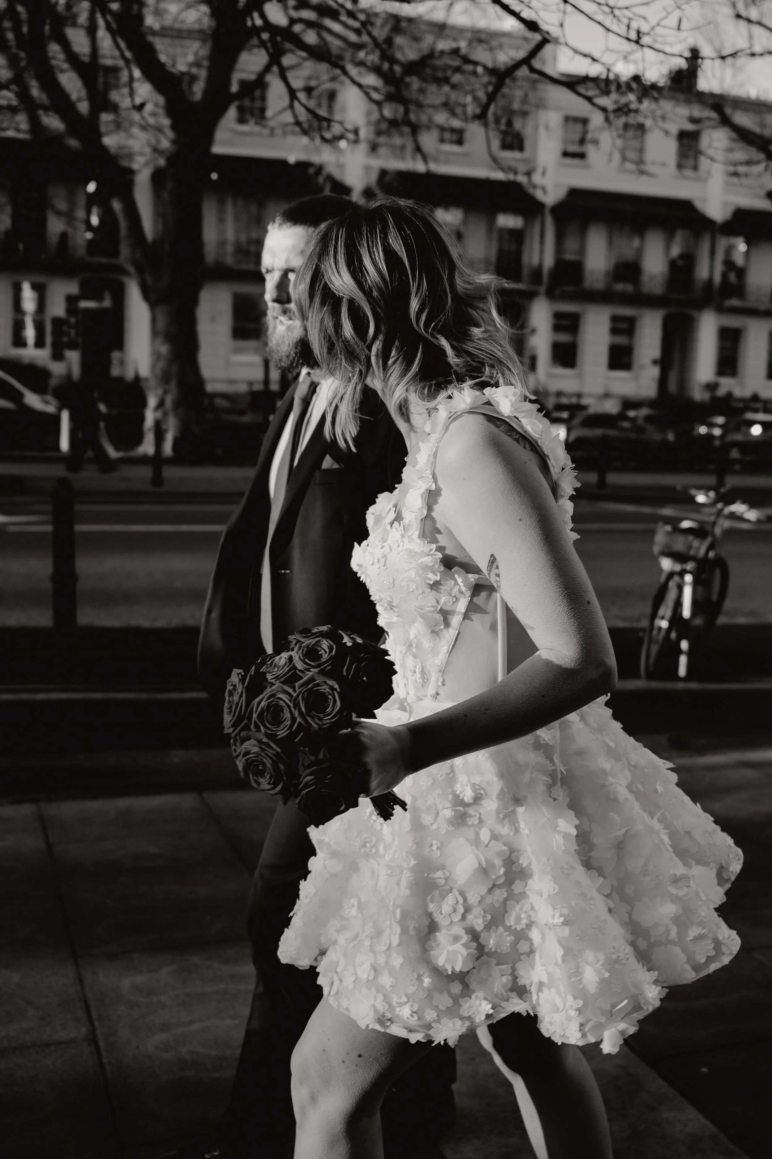 A black and white photo of a bride with shoulder-length wavy hair in a floral wedding dress holding a bouquet of roses, walking next to a bearded man in a dark suit. They're outdoors with trees and buildings in the background.