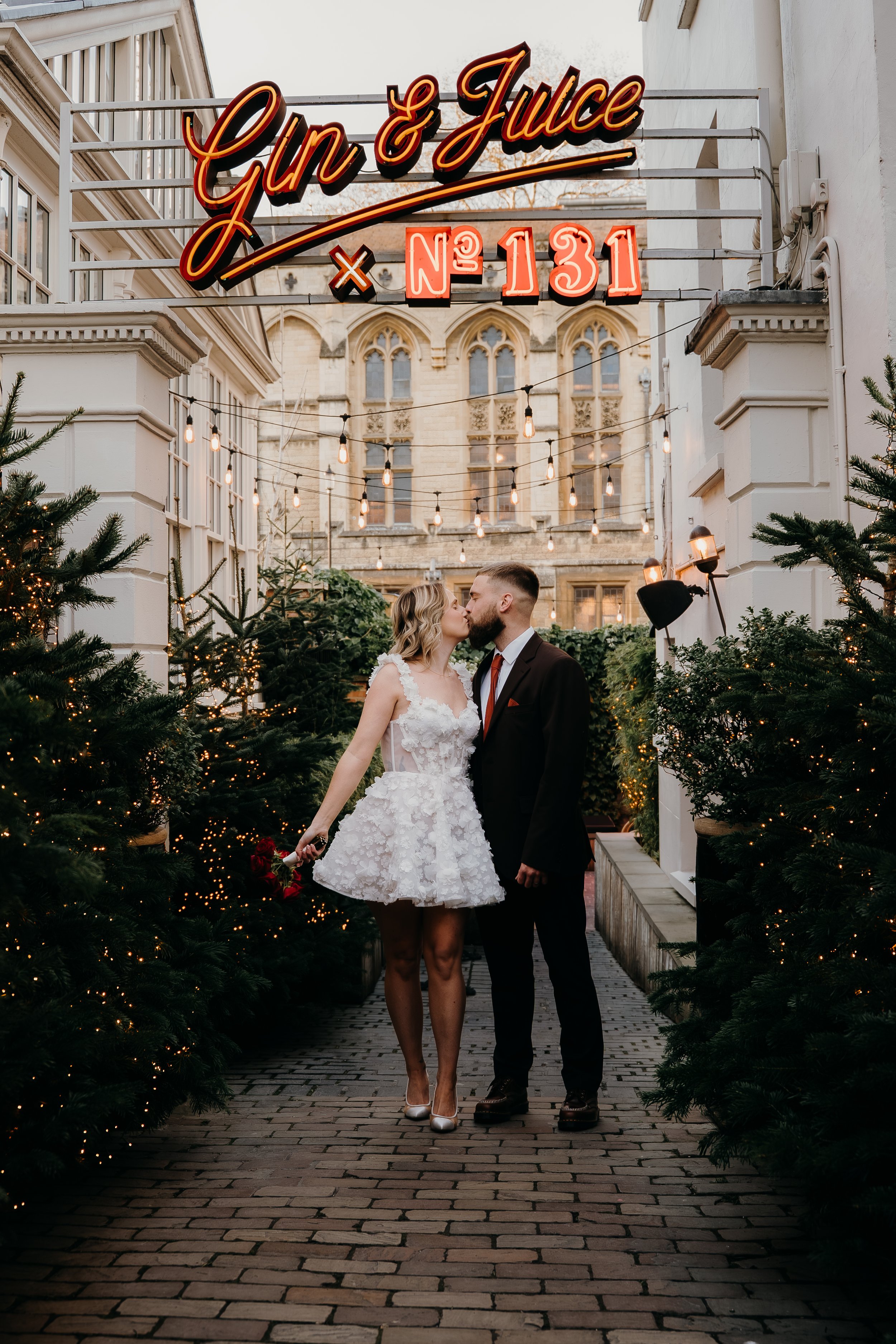 A couple dressed in wedding attire standing nose-to-nose in a festive outdoor setting with Christmas trees and string lights, under a neon sign that says 'Gin & Juice' with the address number 131.