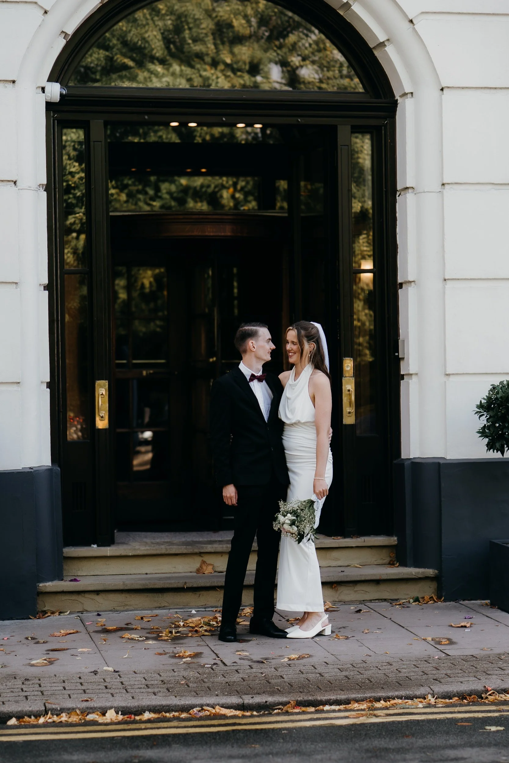 A newlywed couple standing on the steps outside a building, smiling and looking at each other as they pose for a photo. Iris willow photography bar 131 Cheltenham 