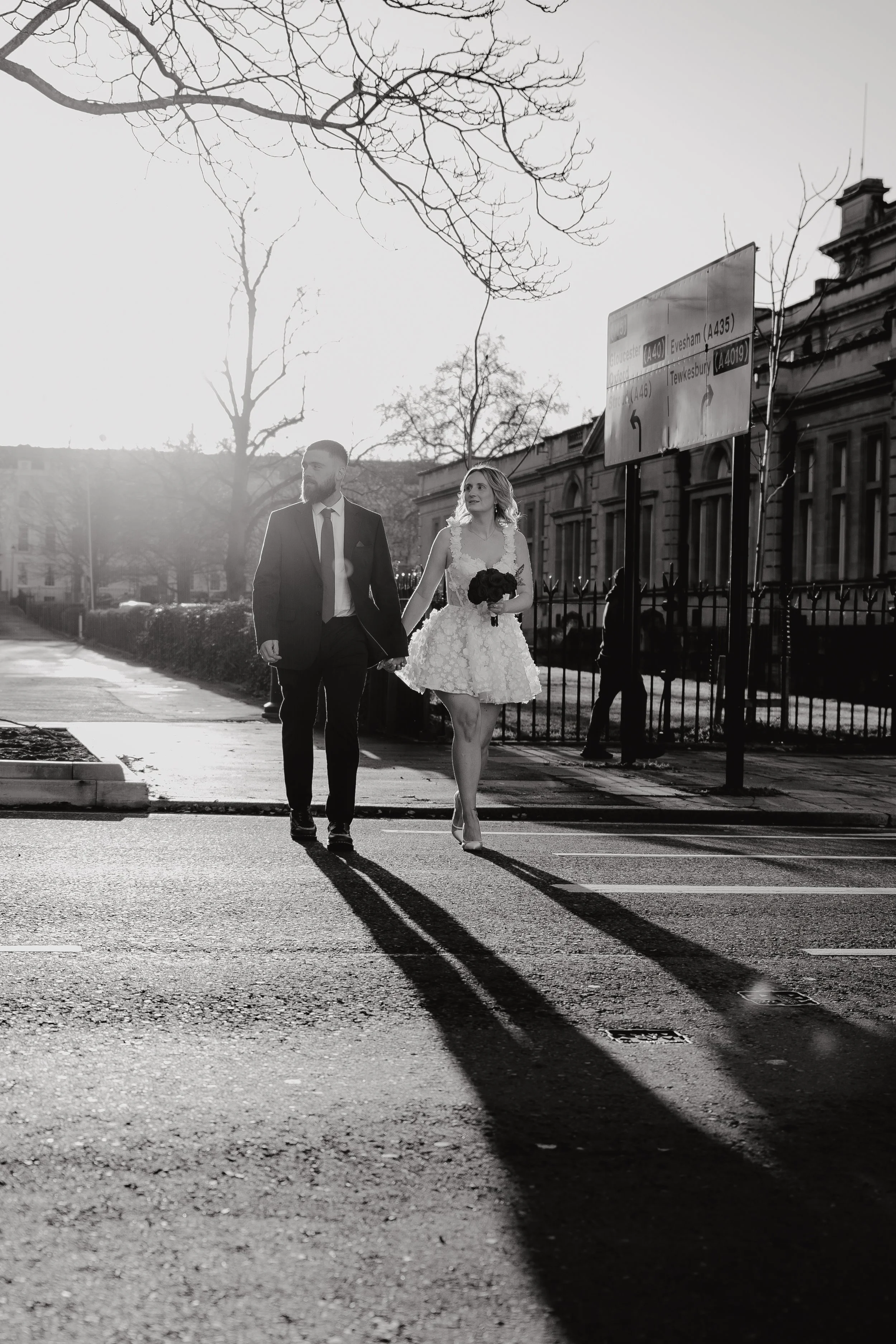 A bride in a wedding dress holding a bouquet and a groom in a suit walking hand in hand on a city street with shadows cast by the setting sun, leafless trees, and a city building in the background.