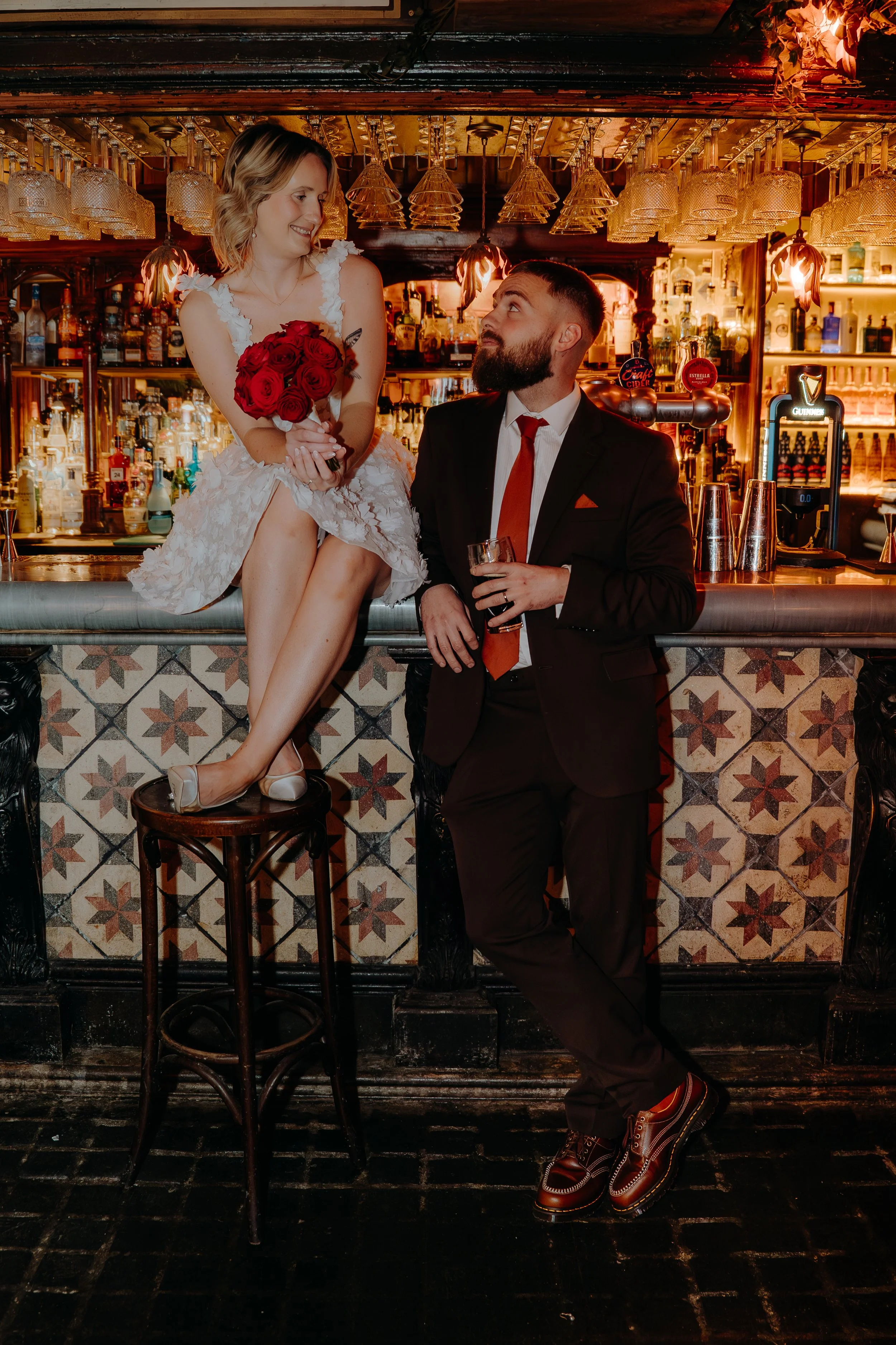 A Bride in a white dress holding a bouquet of red roses, sitting on a stool, smiling at the groom , who is standing next to her in bar 131 Cheltenham. Iris willow photography.