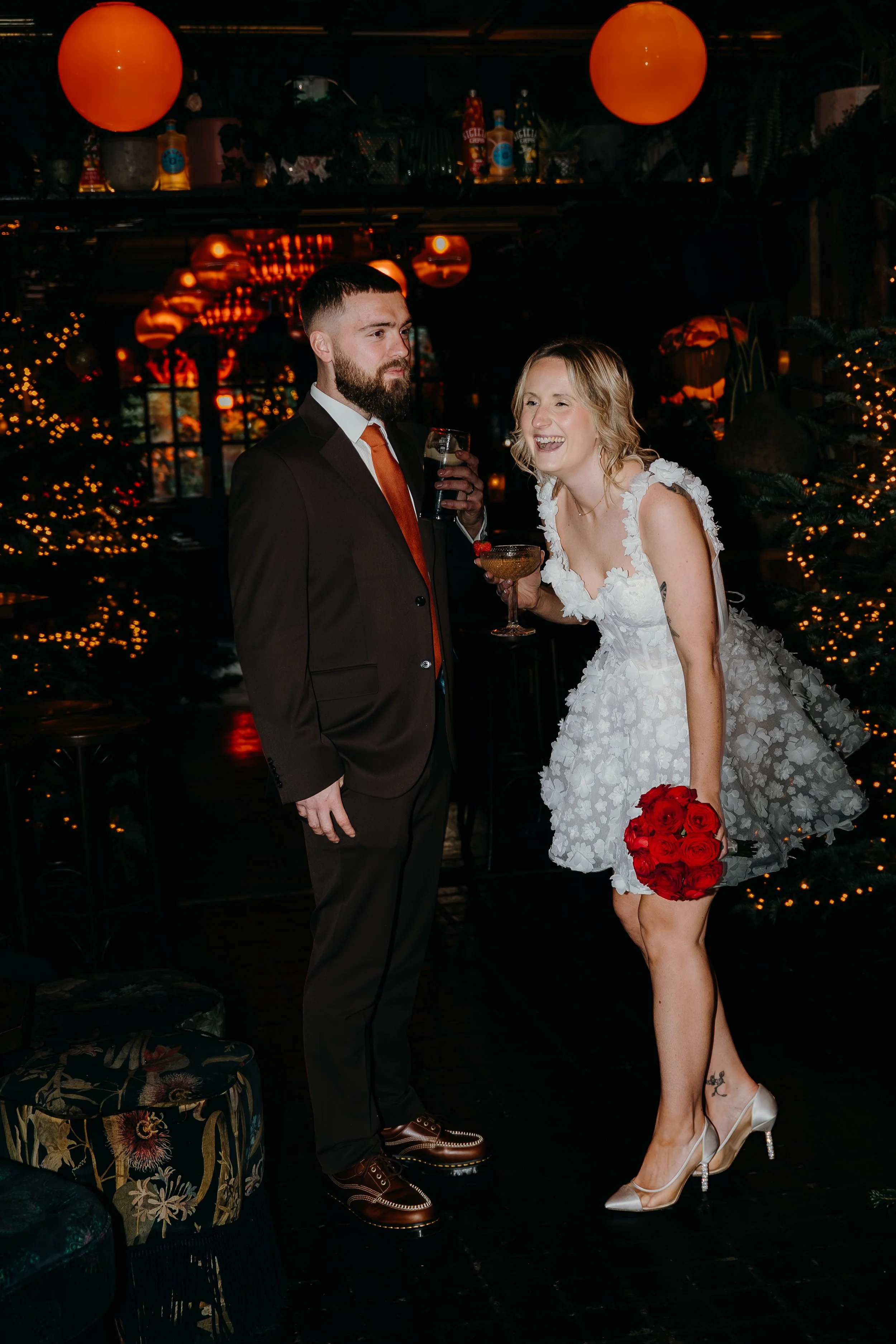 A bride in a white floral dress holding a bouquet of red roses and a groom in a black suit with an orange tie, standing in a dimly lit room decorated with orange lanterns and string lights, sharing a moment during a wedding.