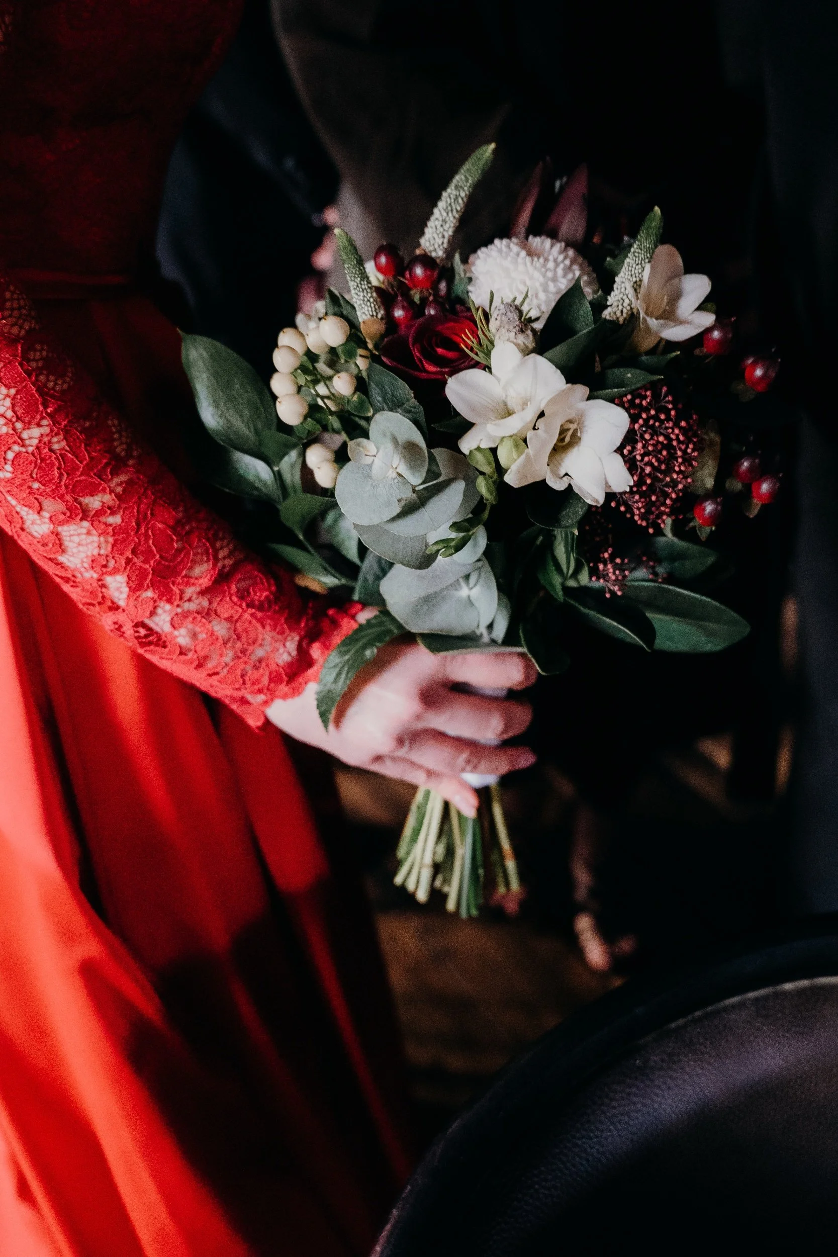 Person in red lace sleeve holding a bouquet of mixed flowers including white, red, and pink blossoms and green foliage.