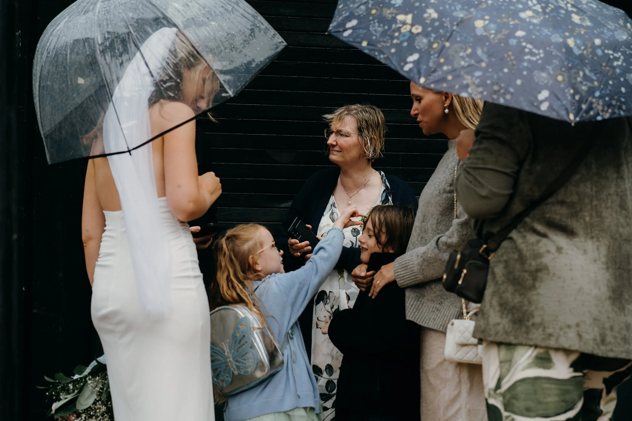 A bride wearing a white dress is receiving a bouquet of flowers from a young girl in a blue jacket, surrounded by women with umbrellas during a rainy outdoor wedding ceremony.