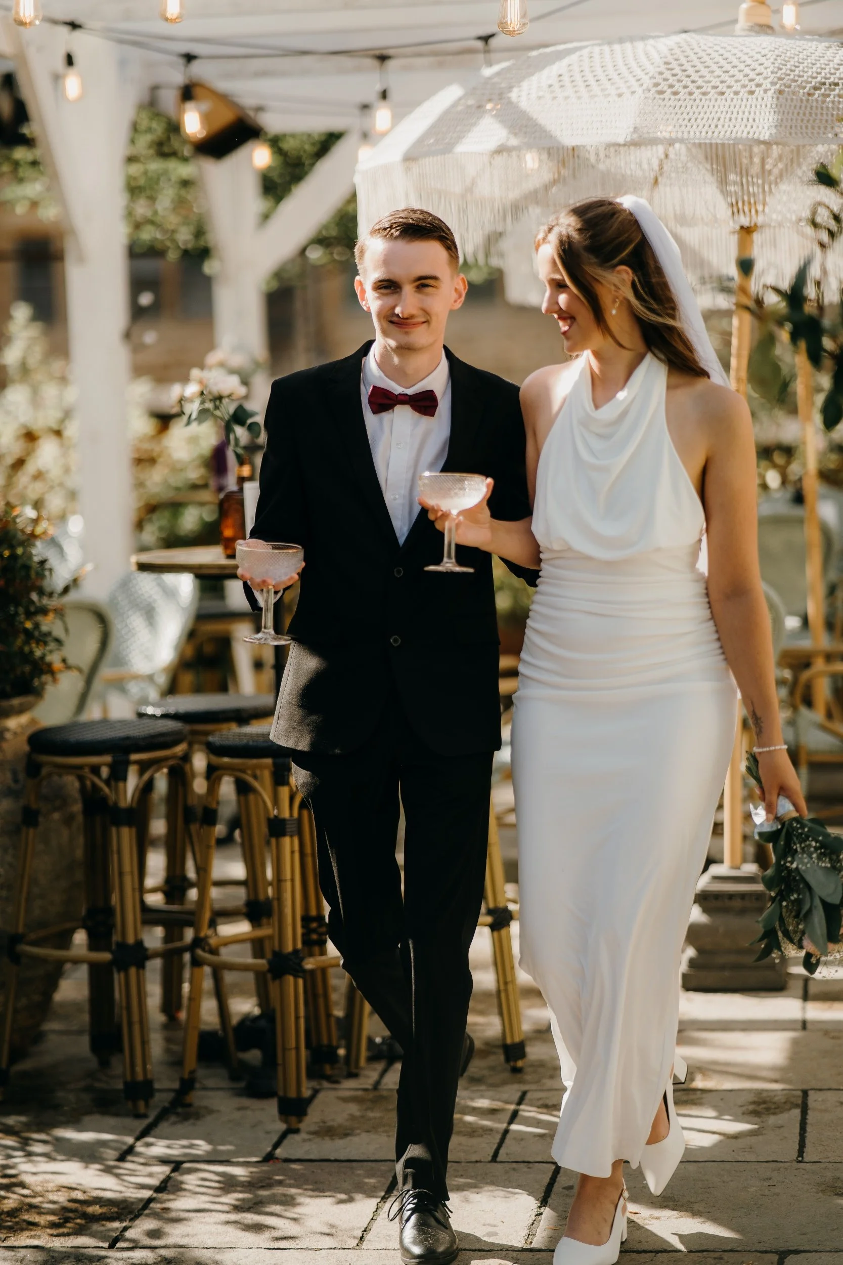 A newly married couple walking together outdoors, holding glasses, under a decorative parasol, with a white wedding dress. Iris willow photography bar 131 Cheltenham 
