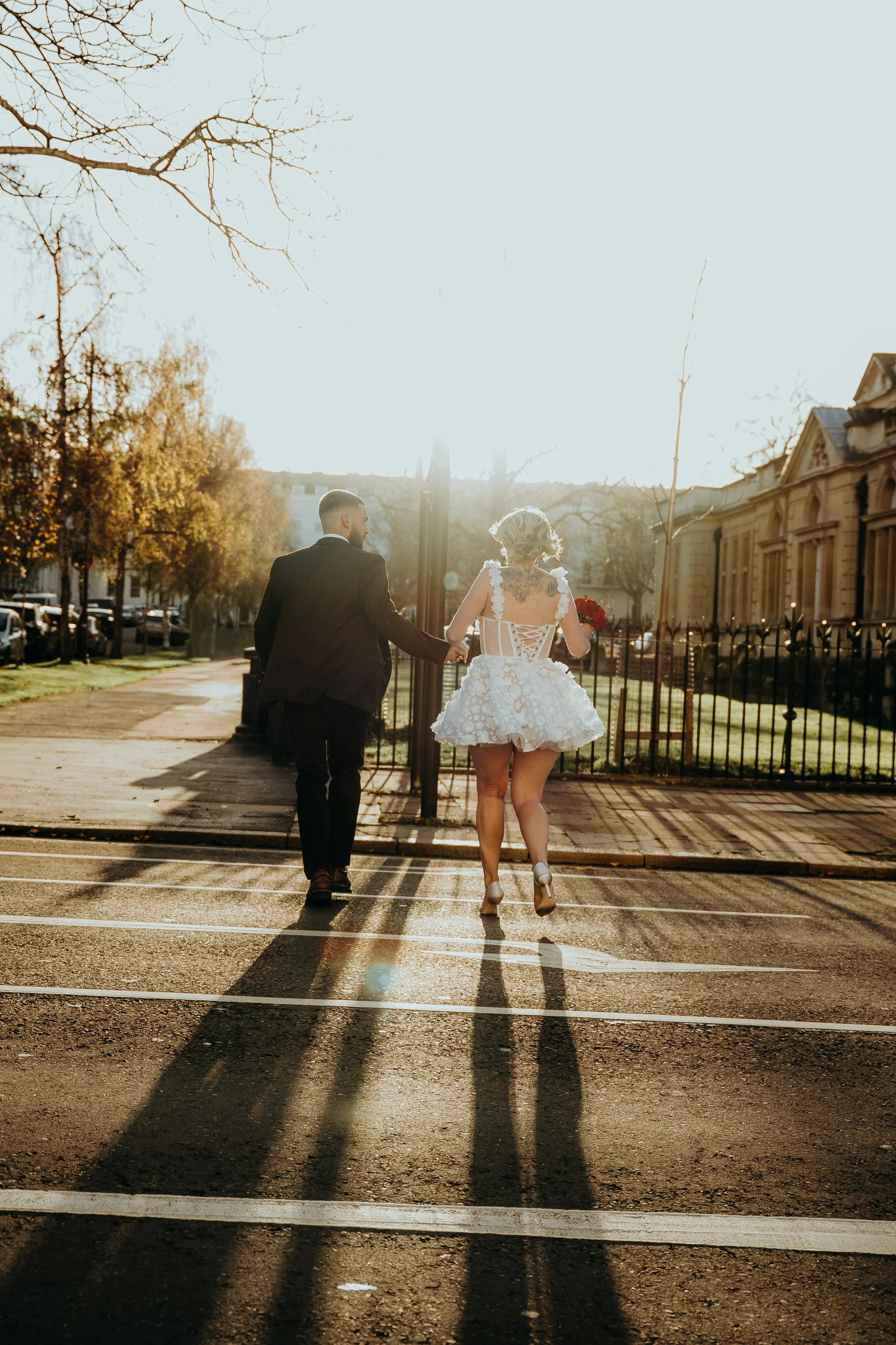 A couple, a man in a black suit and a woman in a white dress, walk hand-in-hand across a crosswalk in a sunny urban setting, with trees and historic buildings in the background.