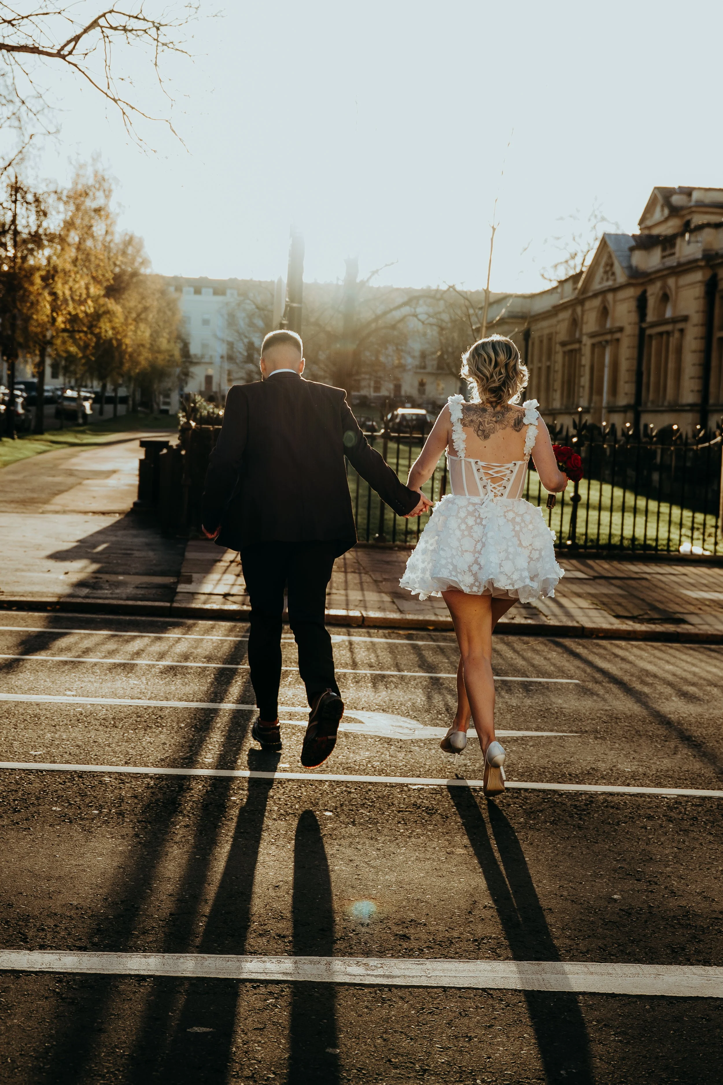 A bride and groom holding hands and walking away on a city street during sunset, casting long shadows. The bride wears a white dress with floral details and high heels, while the groom is dressed in a suit.
