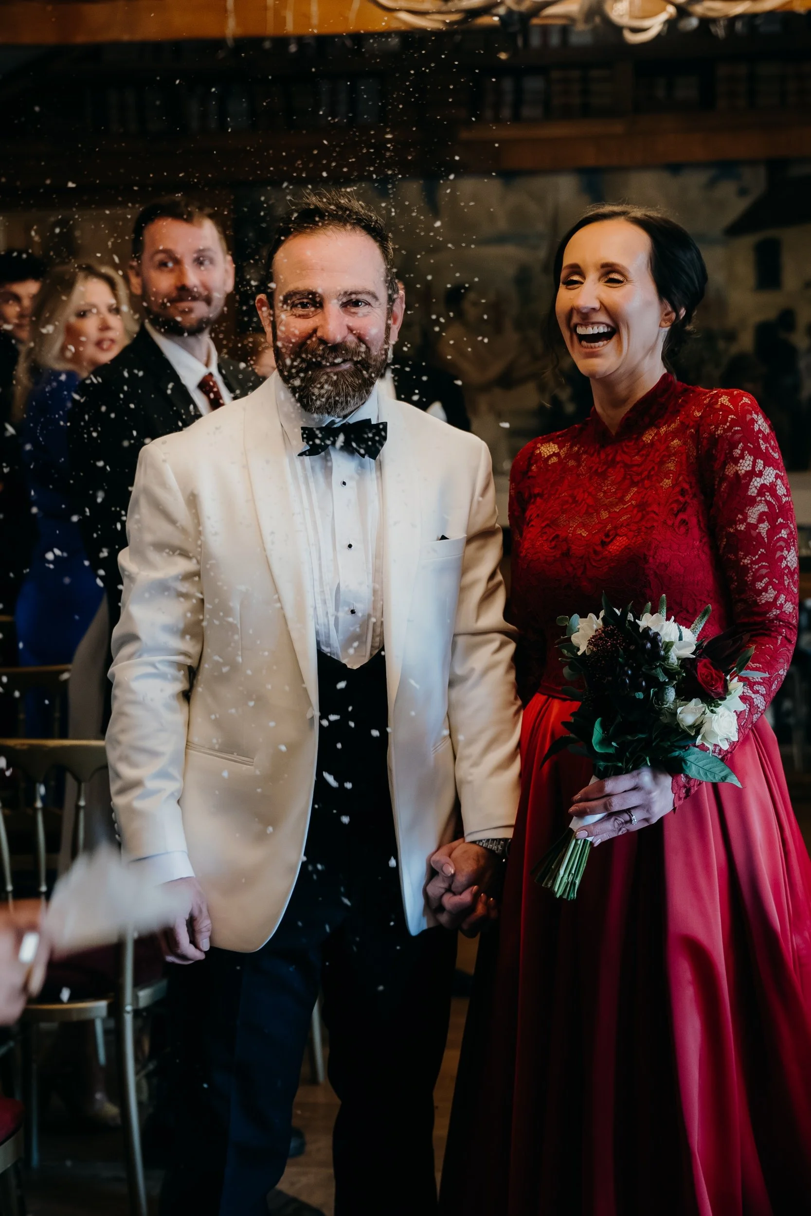 A bride and groom holding hands at their wedding, smiling, with guests in the background. The bride wears a red lace dress and holds a bouquet, while the groom is in a white tuxedo with a black bow tie. Confetti is falling around them.